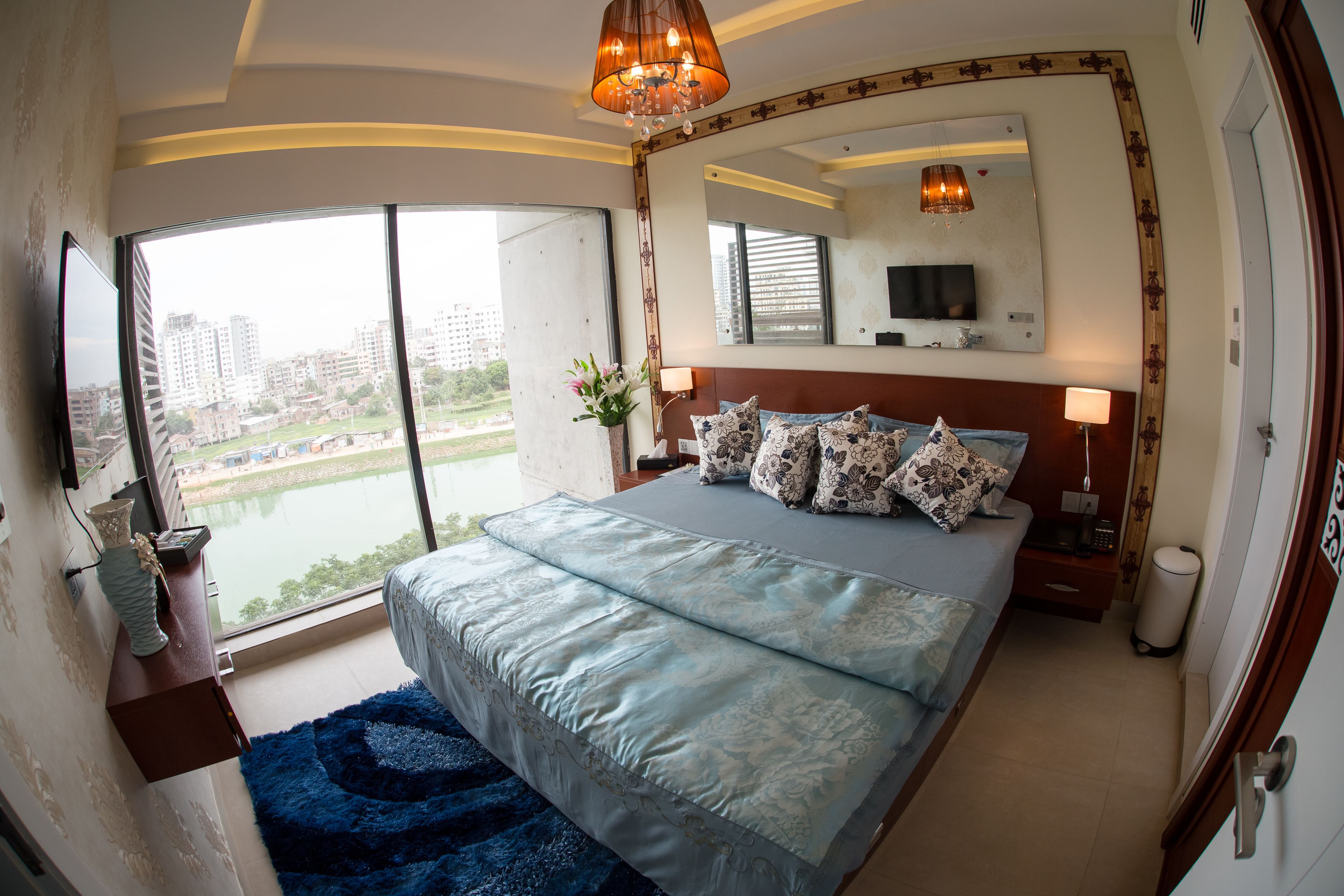 A bedroom with a large bed, a TV, a mirror, and a view of a cityscape through a large window.