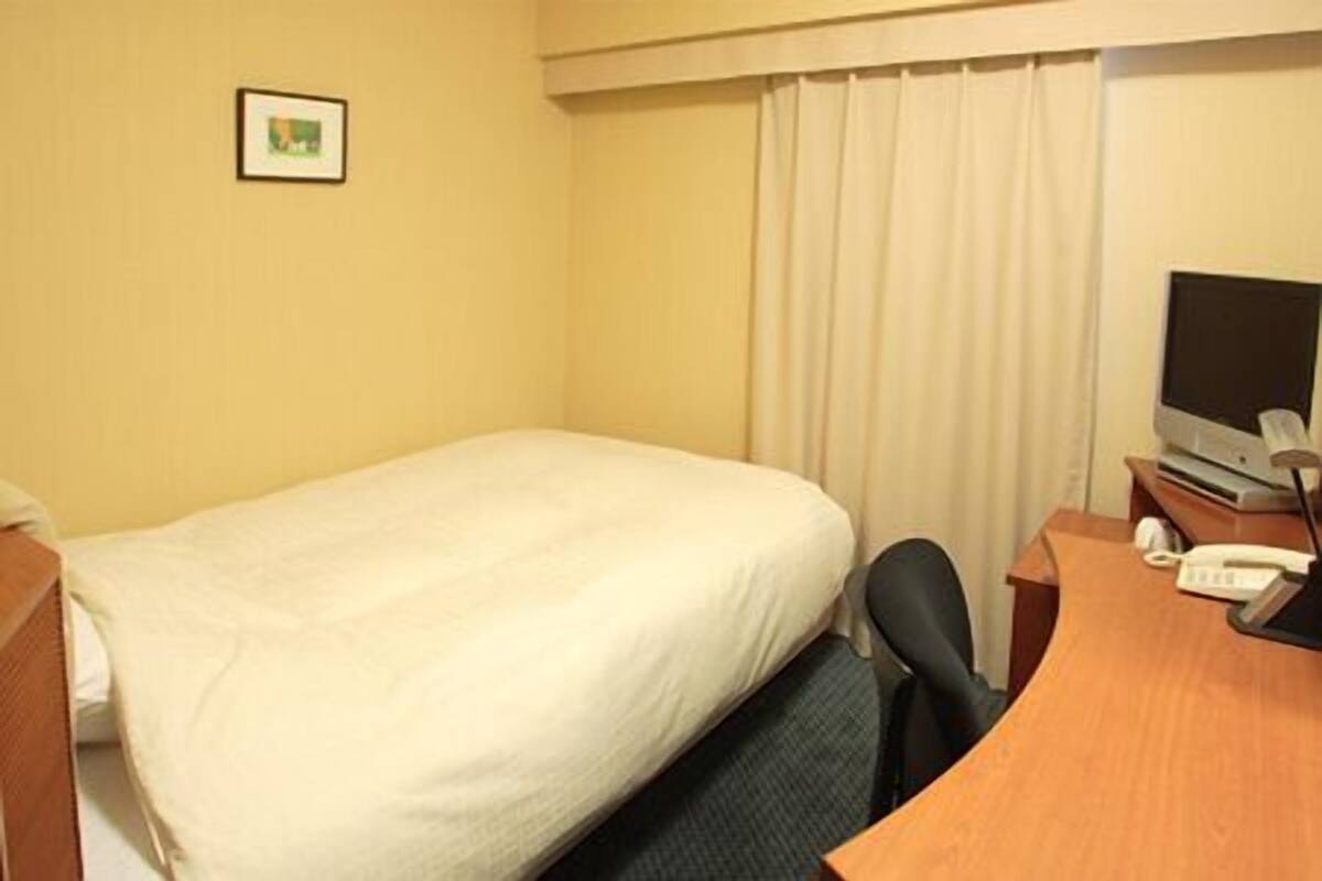 Quarto de hotel com uma cama de solteiro, uma escrivaninha com computador e um telefone.