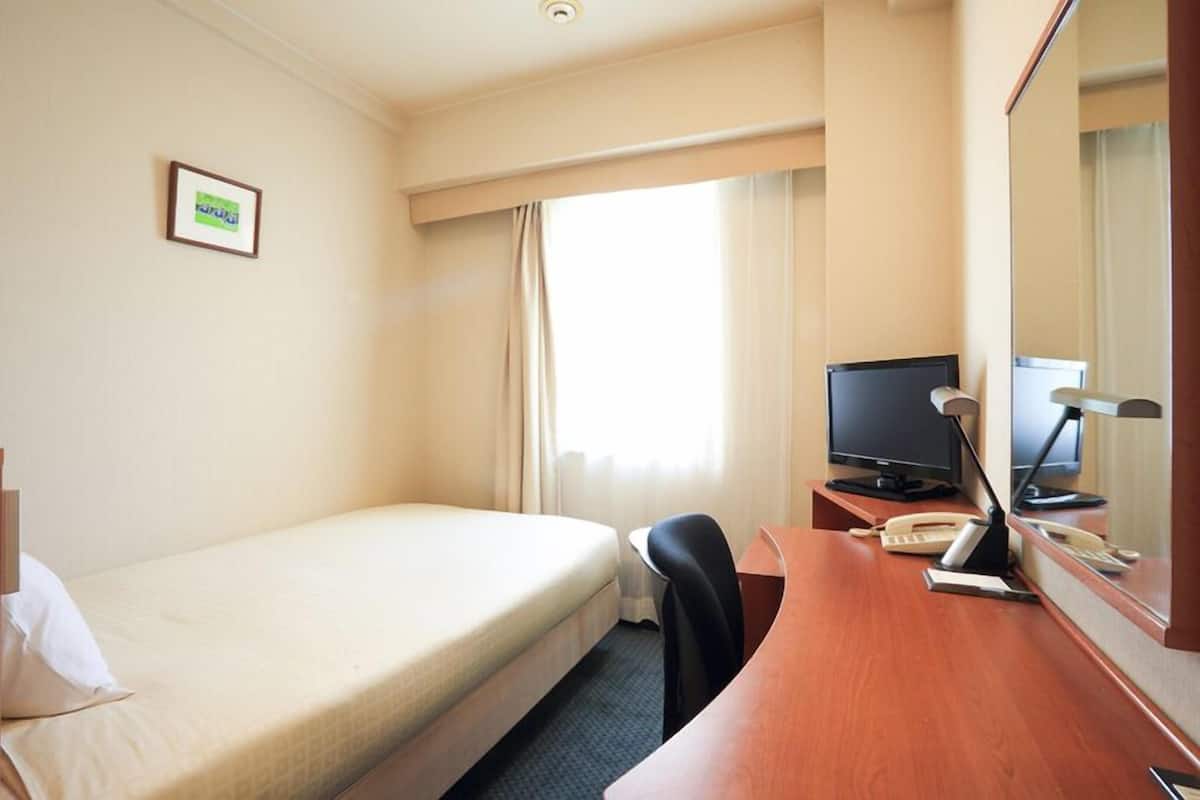 Quarto de hotel com uma cama de solteiro, uma escrivaninha com computador, telefone e televisão.