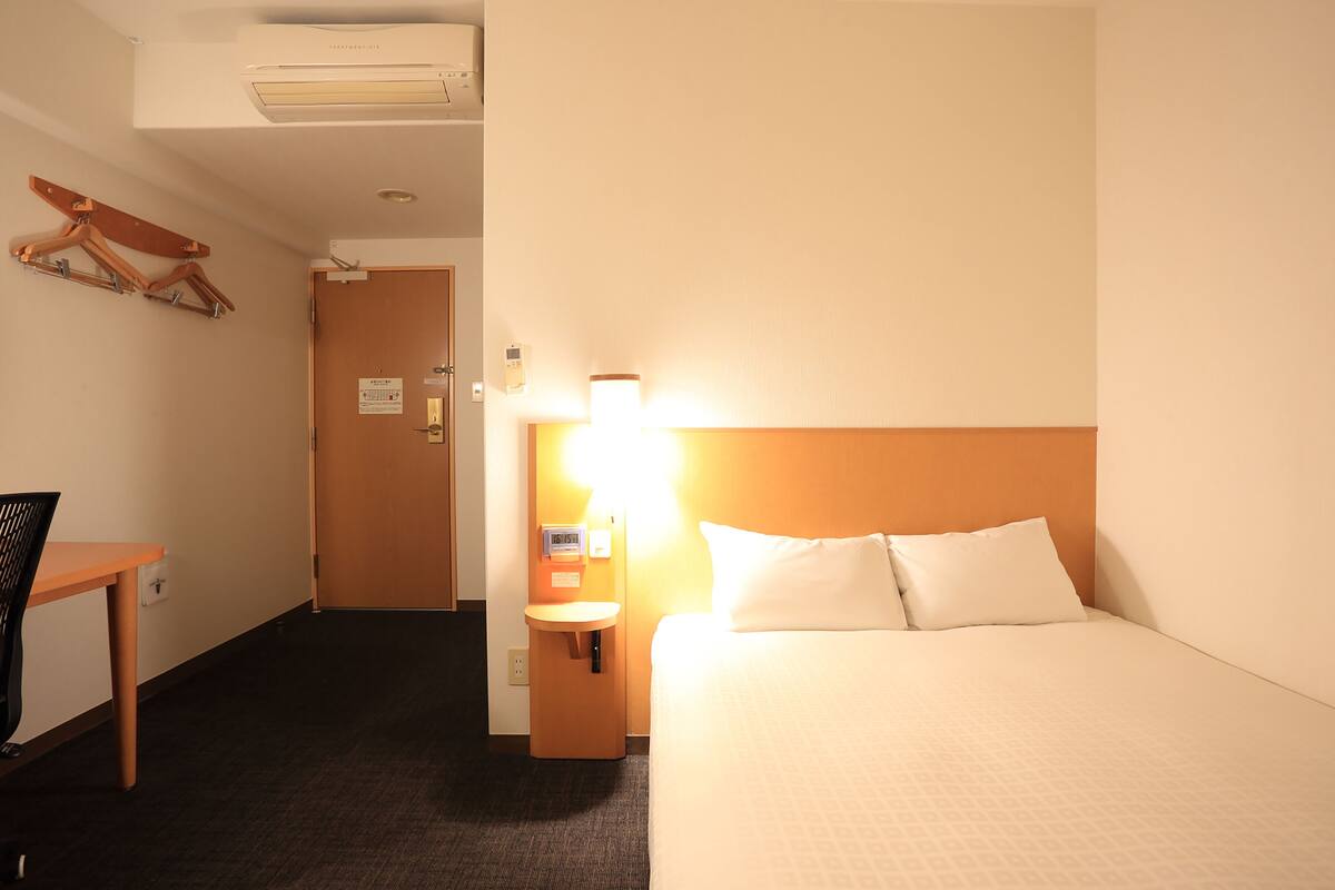 Quarto de hotel com cama, escrivaninha, cadeira, aquecedor na parede e porta.