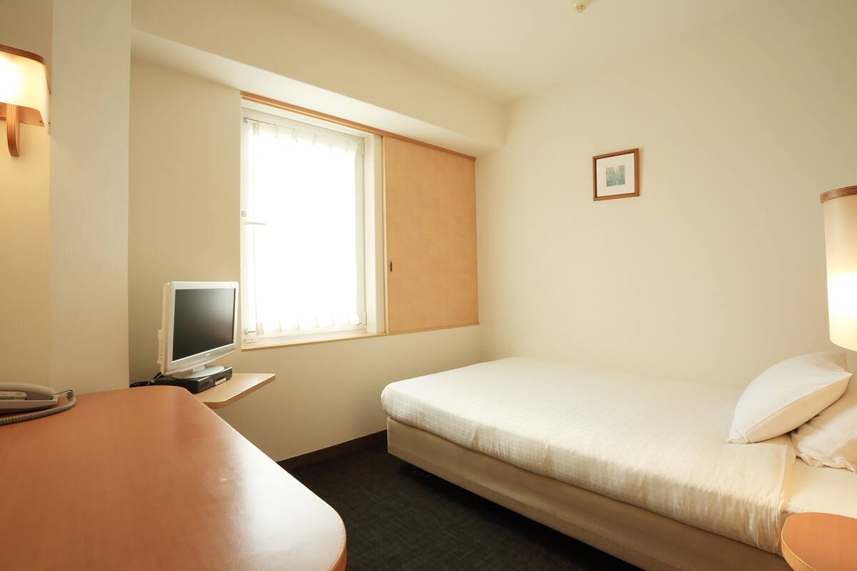 Quarto de hotel com uma cama, uma escrivaninha, televisão e uma janela com persianas.