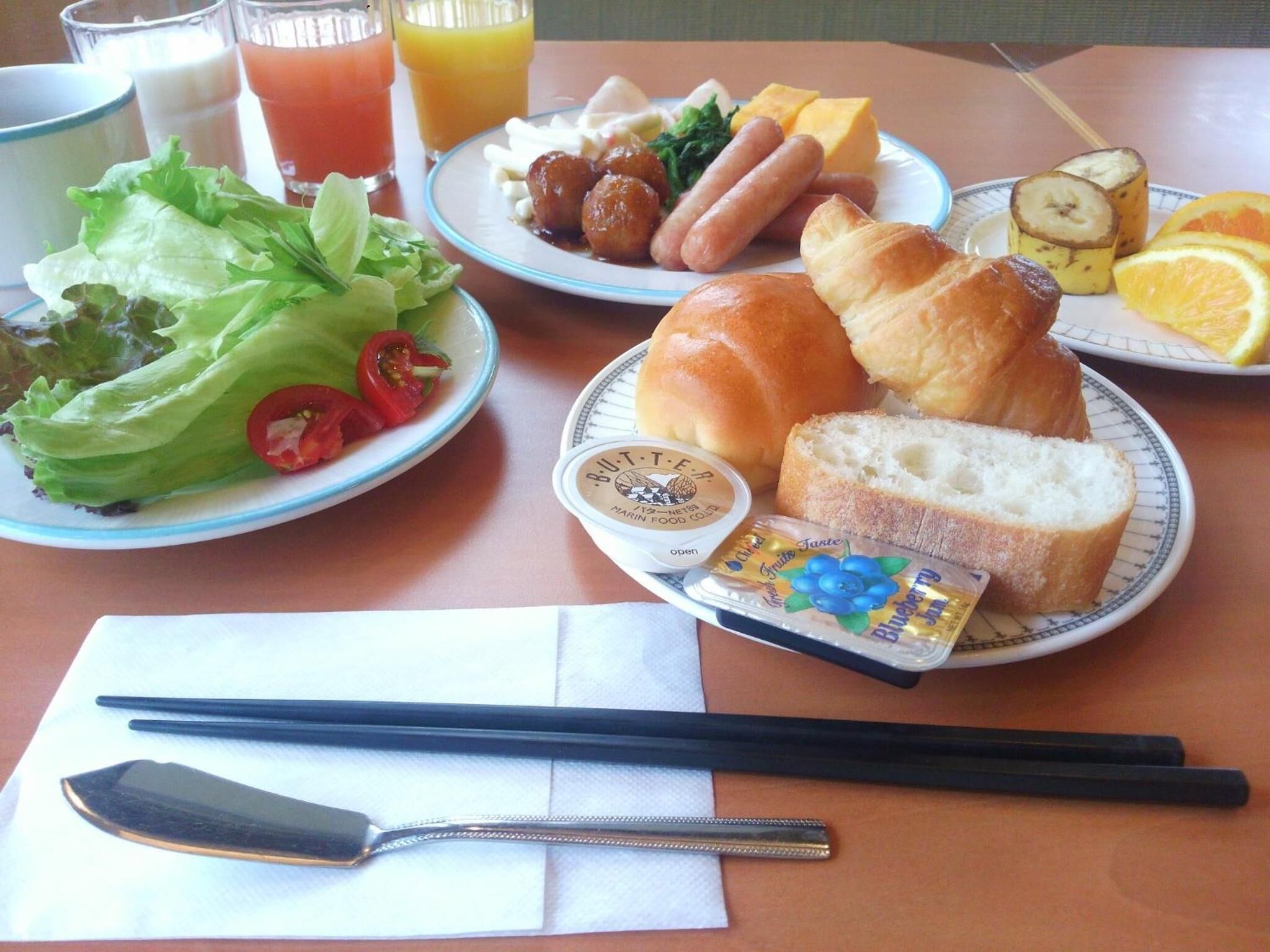 Une table est dressée avec des assiettes remplies de nourriture, notamment des hot-dogs, du pain et une salade. On y trouve également des boissons, une serviette en papier, ainsi que des baguettes et une cuillère.