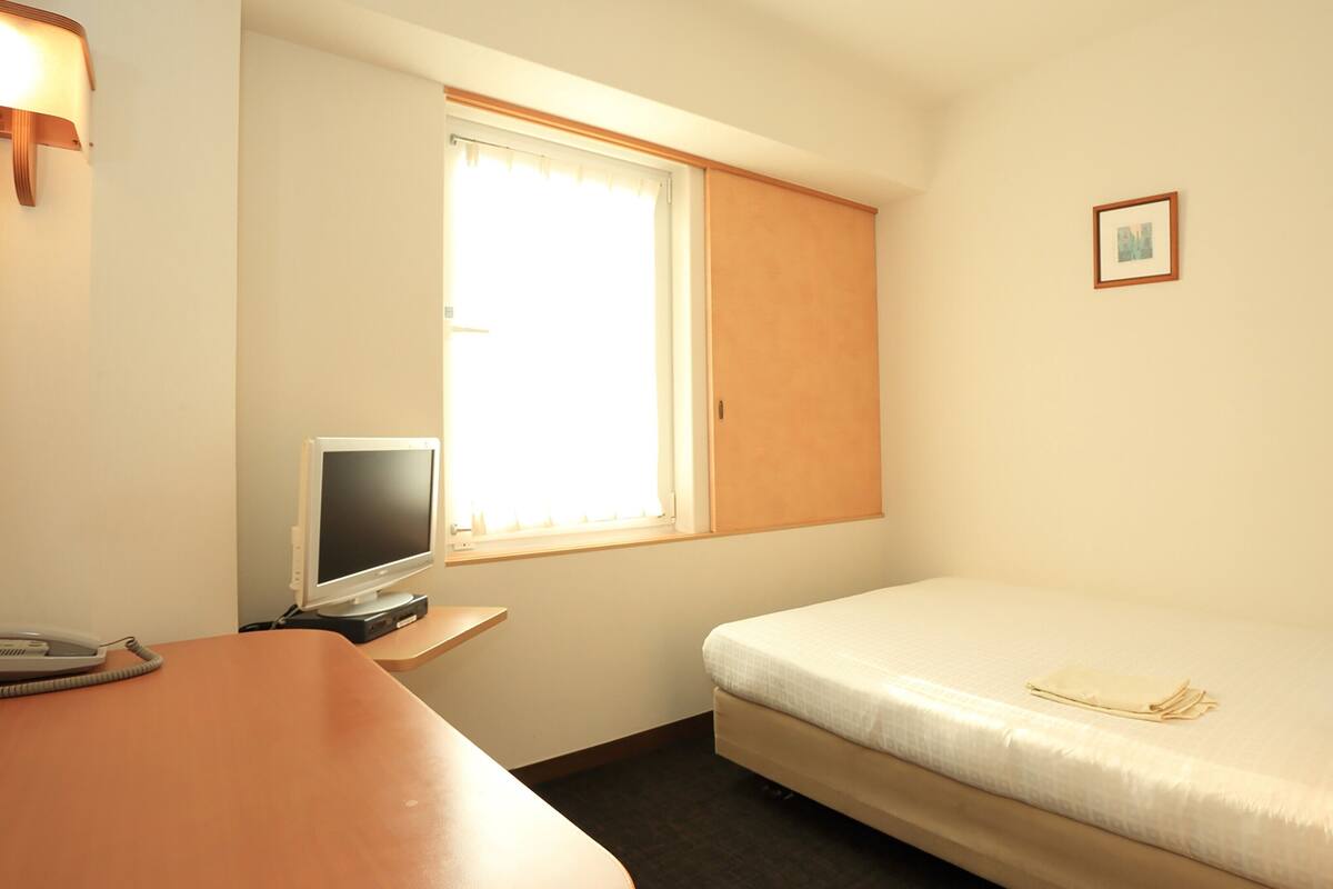 Quarto pequeno com uma cama de solteiro, uma mesa, televisão, janela com persiana e um quadro na parede.