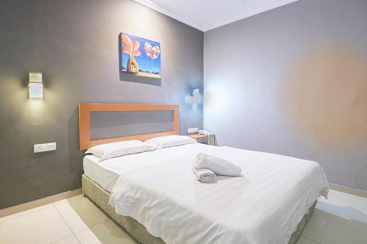 A neatly made bed with white linens and a rolled towel, a wooden headboard, a nightstand with a lamp, and a painting of a tree and hot air balloons on the wall.