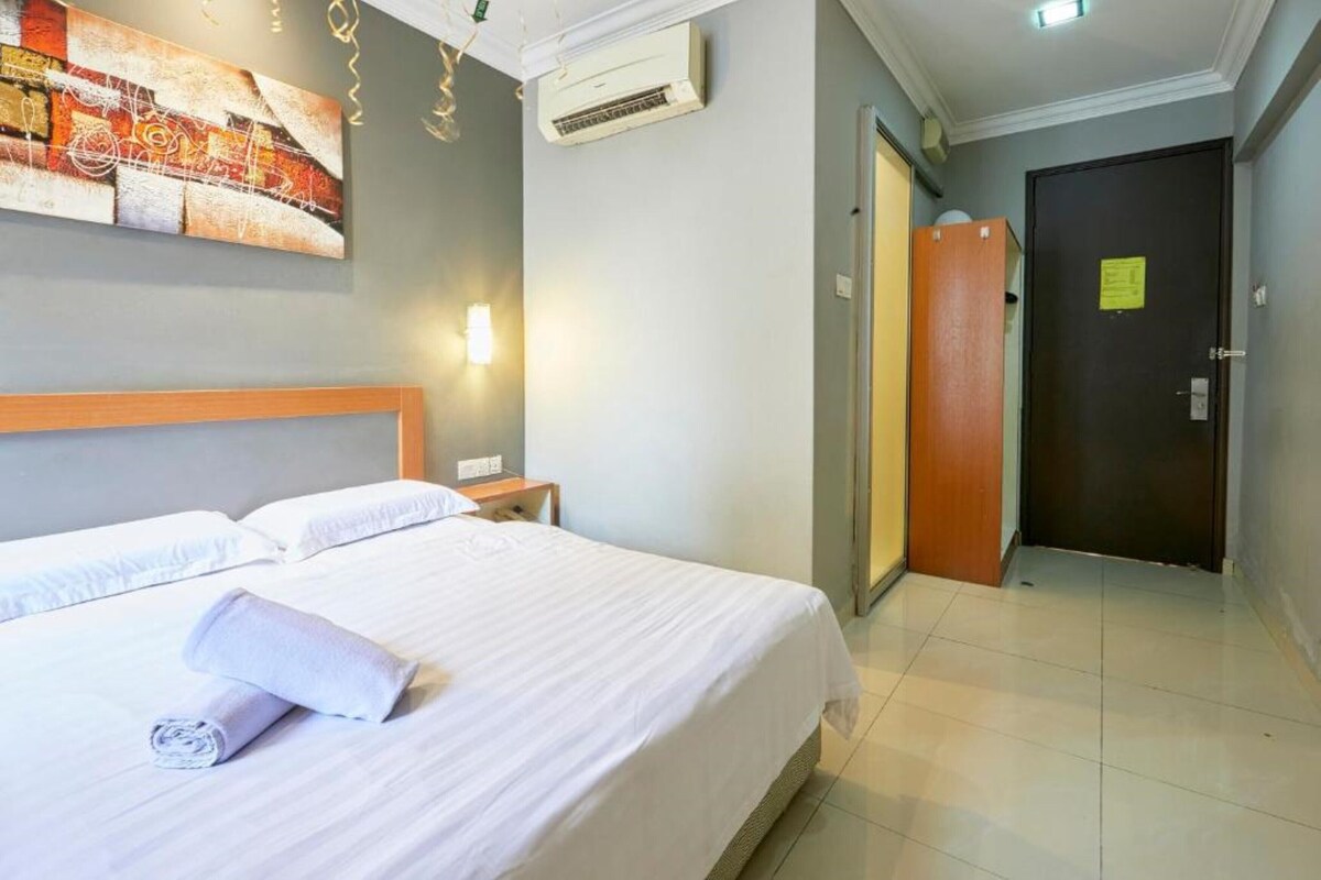 A hotel room with a neatly made bed, a wall-mounted air conditioner, a door with a sign, and a painting on the wall.