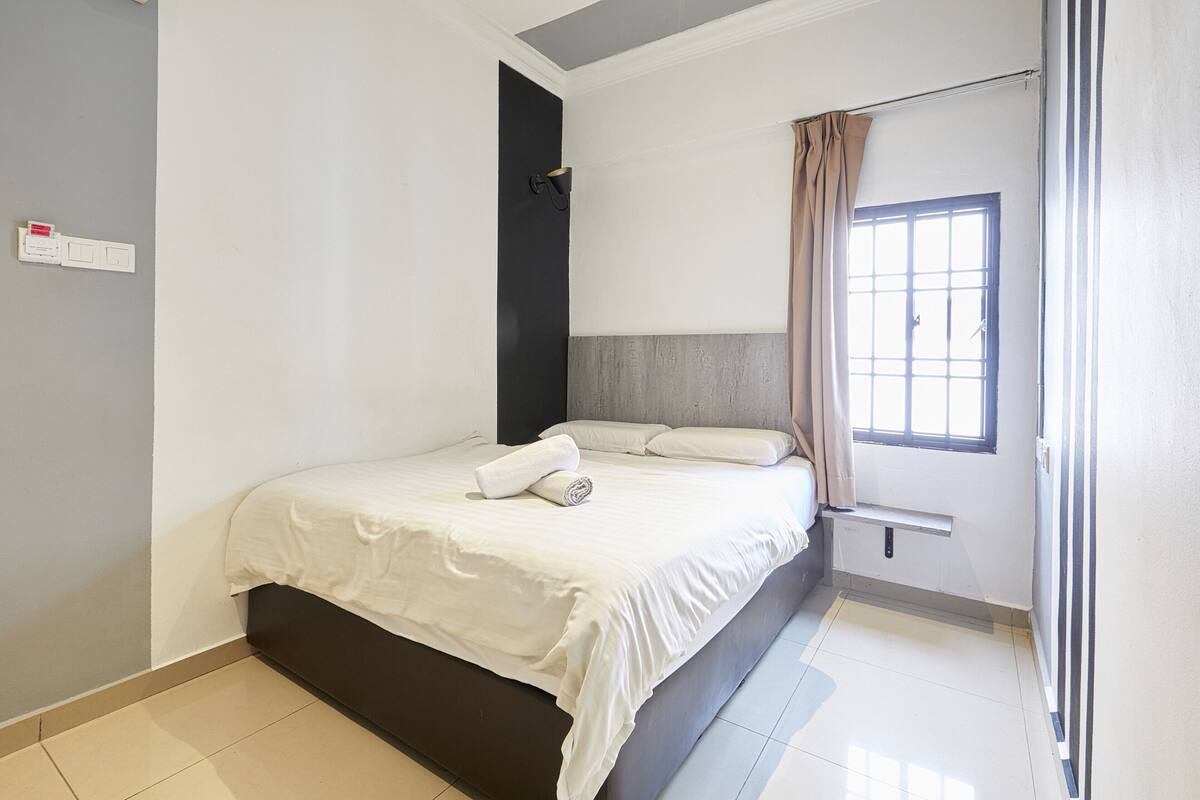 Une chambre à coucher avec un lit, une fenêtre avec des rideaux, une lampe fixée au mur et une prise de courant.