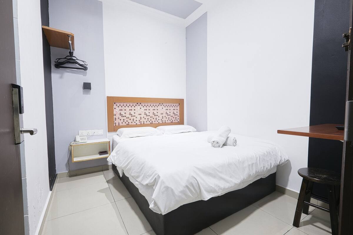 Une chambre d’hôtel bien rangée, avec un lit, une table de chevet et un petit bureau avec une chaise.