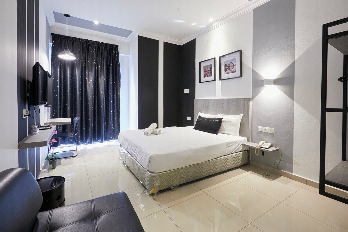 A modern hotel room with a large bed, a desk with a chair, a small table with a plant, and a mirror on the wall.