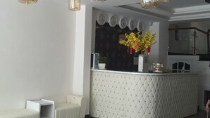 Un área de recepción con un mostrador blanco con textura, una zona de estar con un sofá blanco y una planta decorativa con flores amarillas.