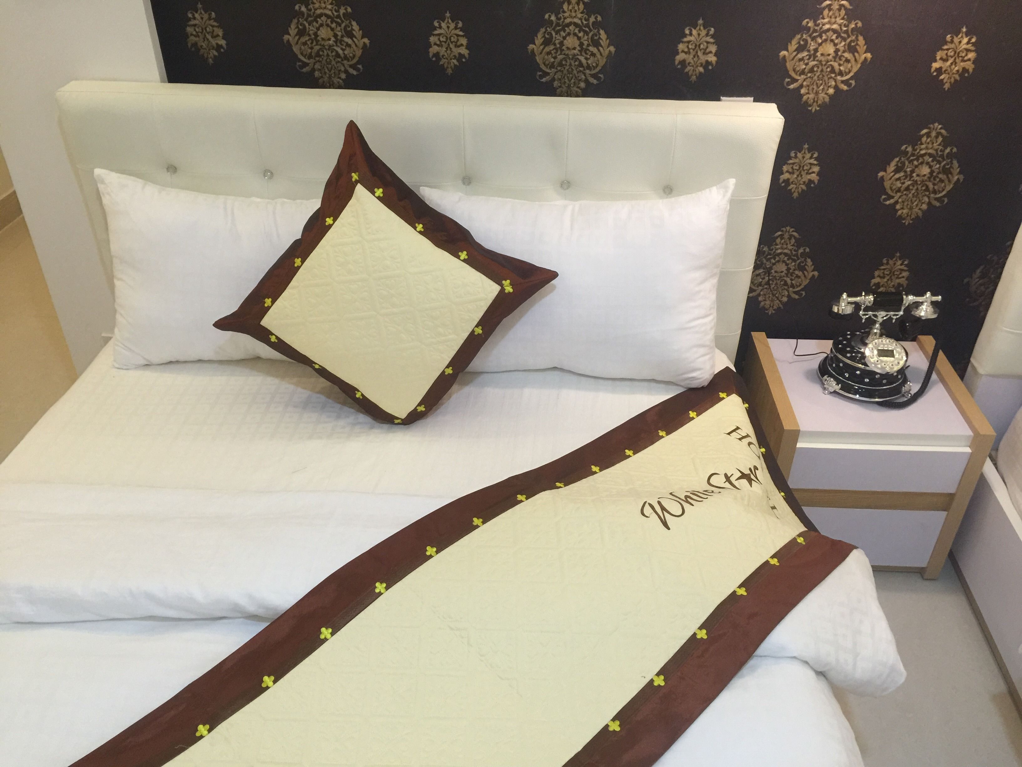 A neatly made bed with a white headboard and a decorative pillow. A bedside table with a telephone and a small lamp is visible.
