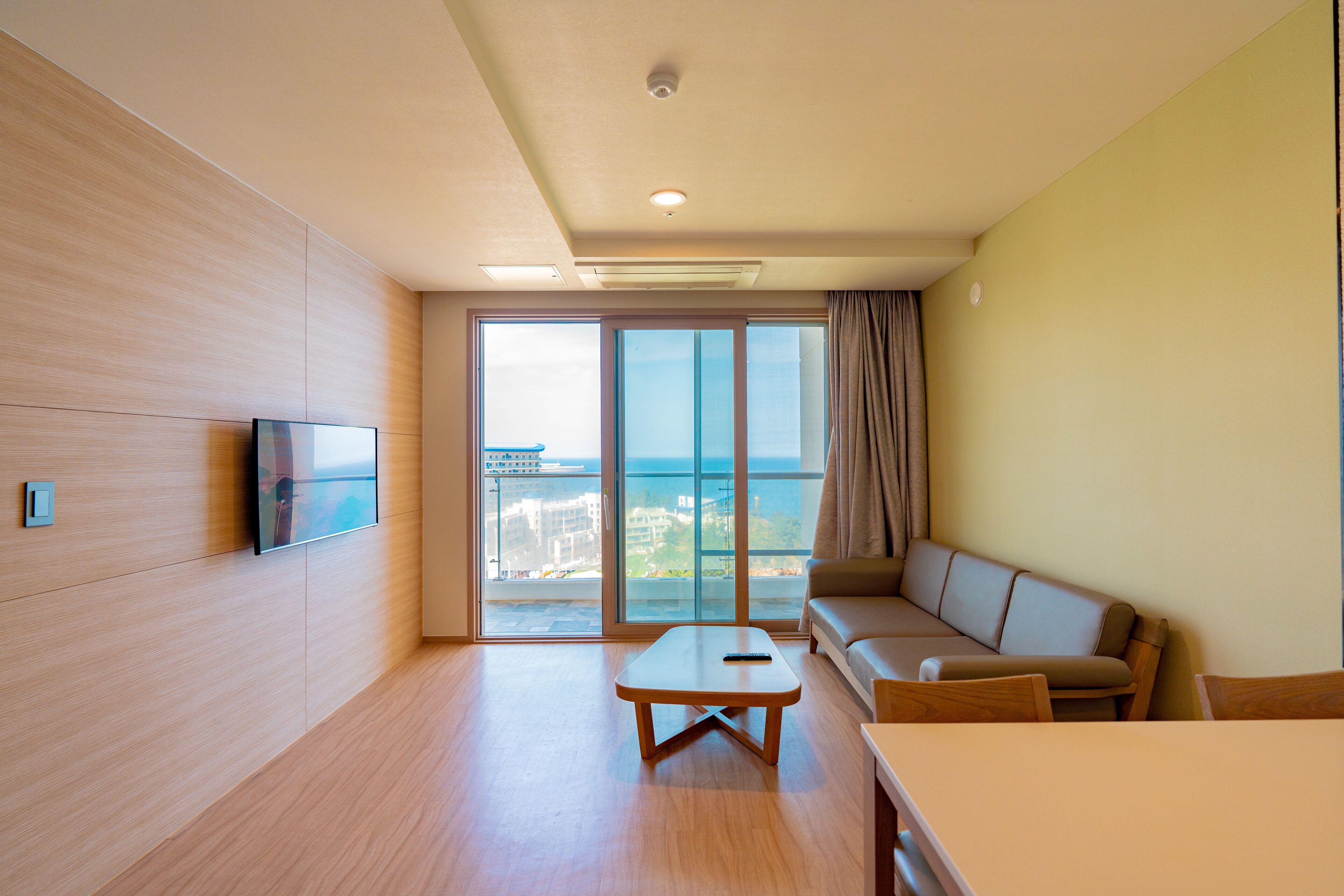 A modern living room with a flat-screen TV, a sofa, a coffee table, and a balcony with a view.