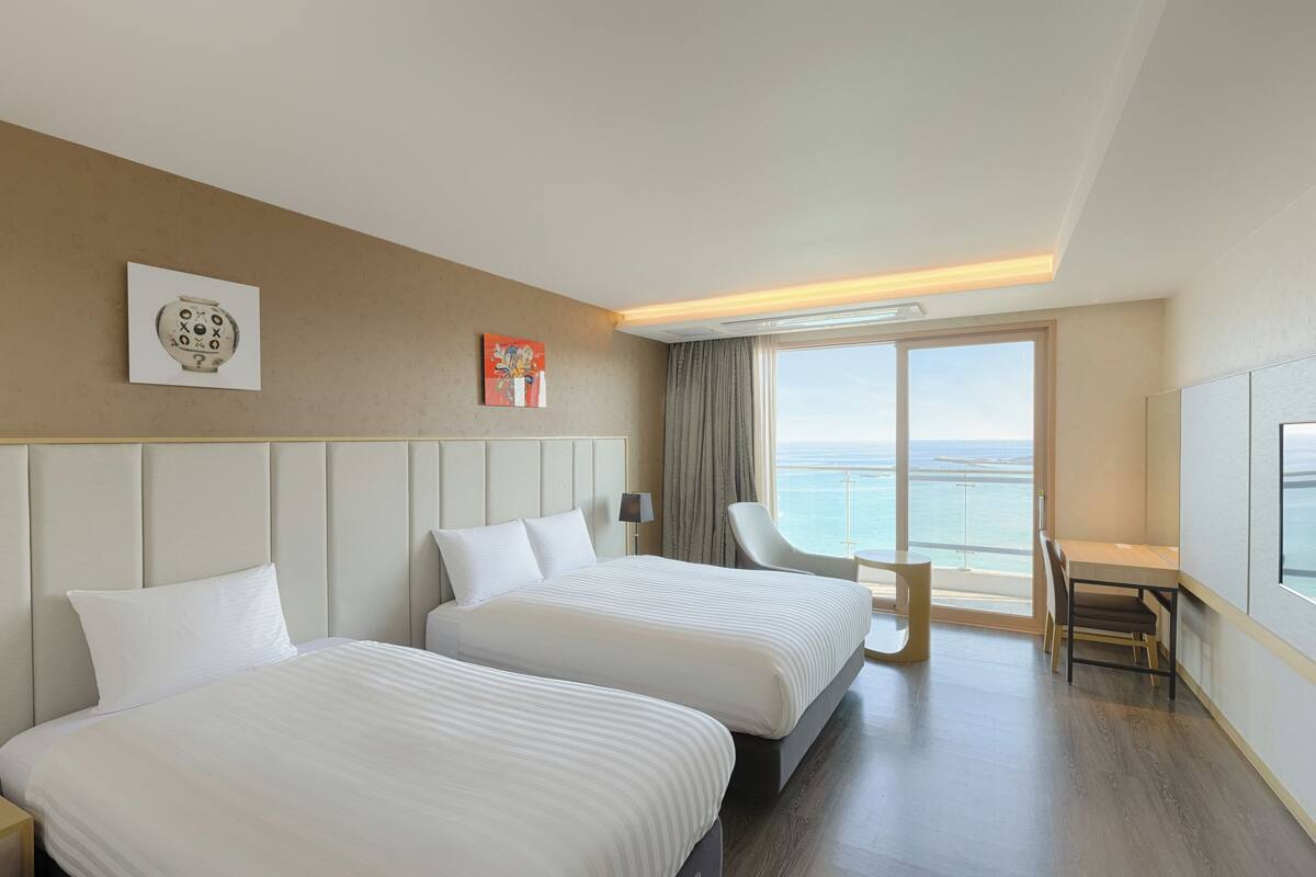 A hotel room with two beds, a desk, a chair, and a large window with a view of the sea.