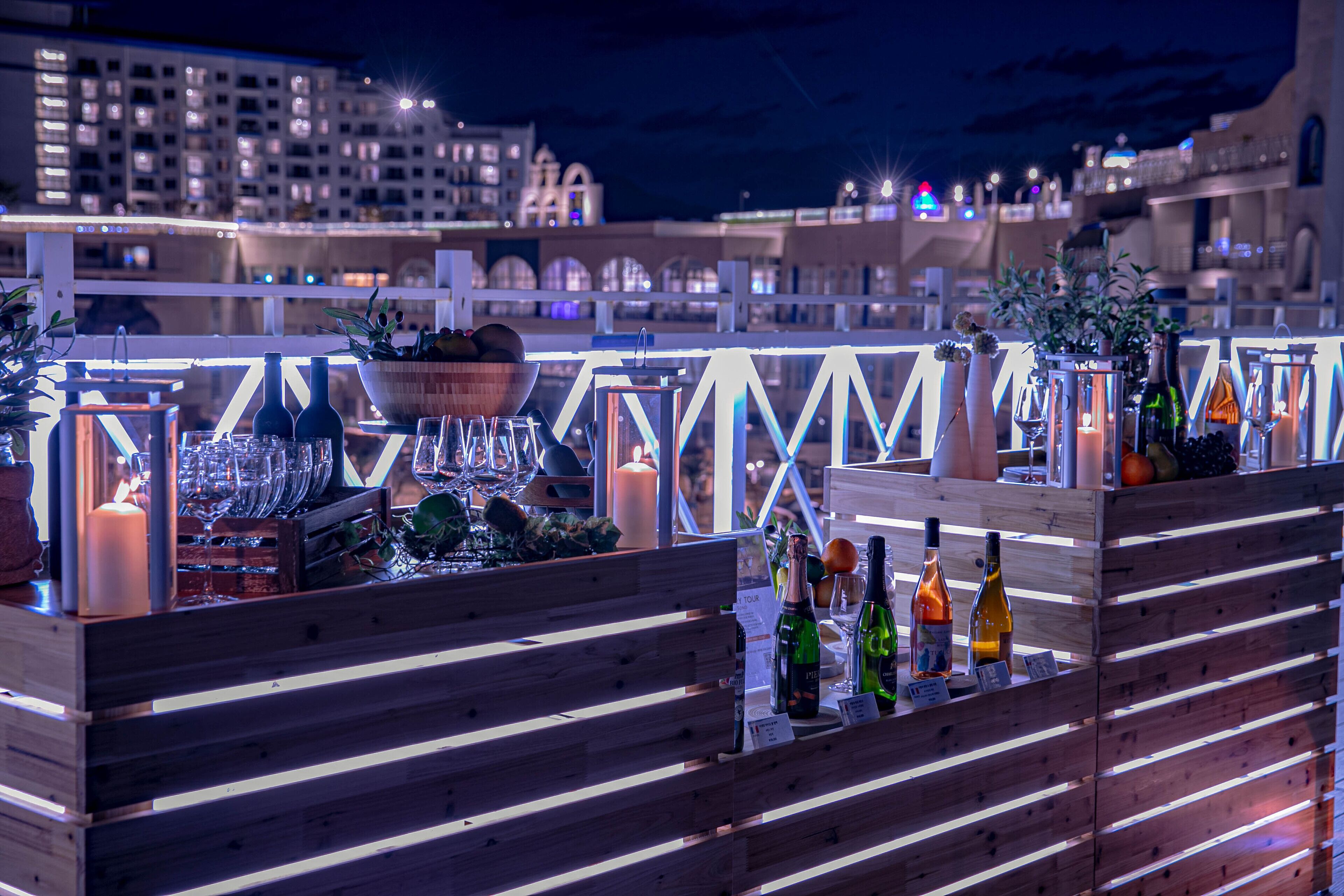 A rooftop bar with wooden seating, bottles, glasses, and a candle.