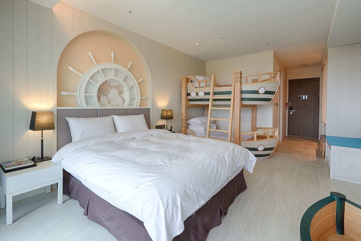 A bedroom with a large bed, a bunk bed, a wooden ship's wheel decoration, a nightstand, and a desk with a lamp.