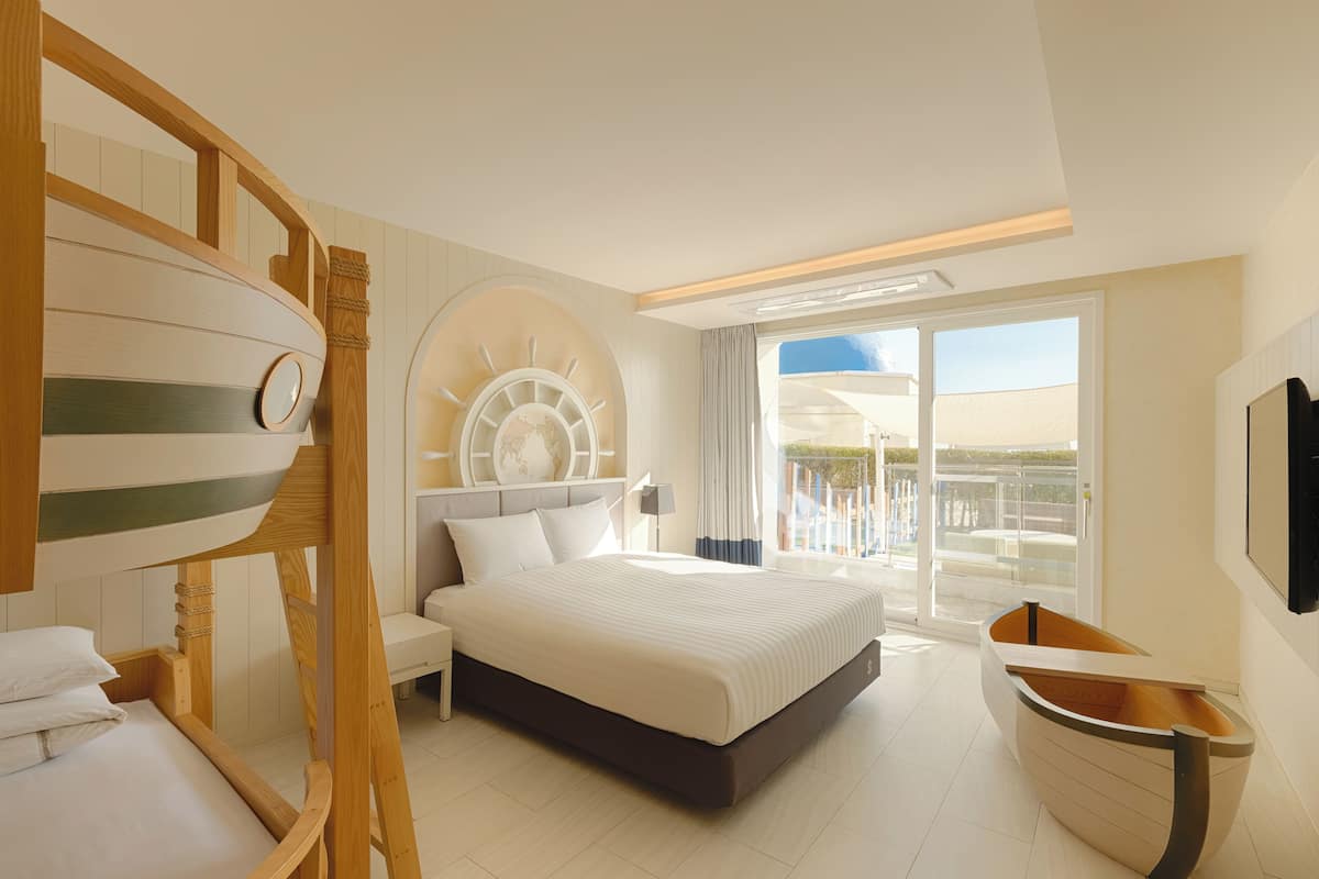 A modern hotel room with a large bed, a wooden boat-shaped side table, a round wooden headboard, and a balcony with a view.
