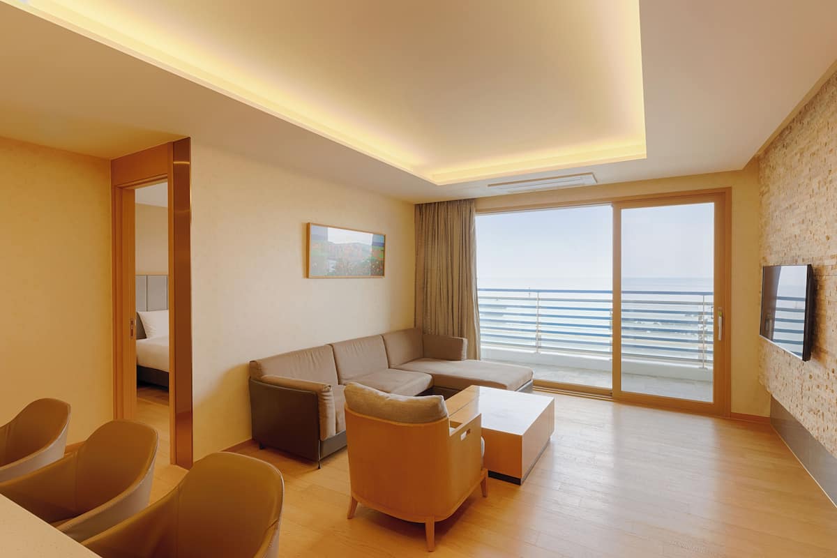 A modern hotel room with a large window offering a view of the sea, a sofa, a coffee table, and a TV mounted on the wall.