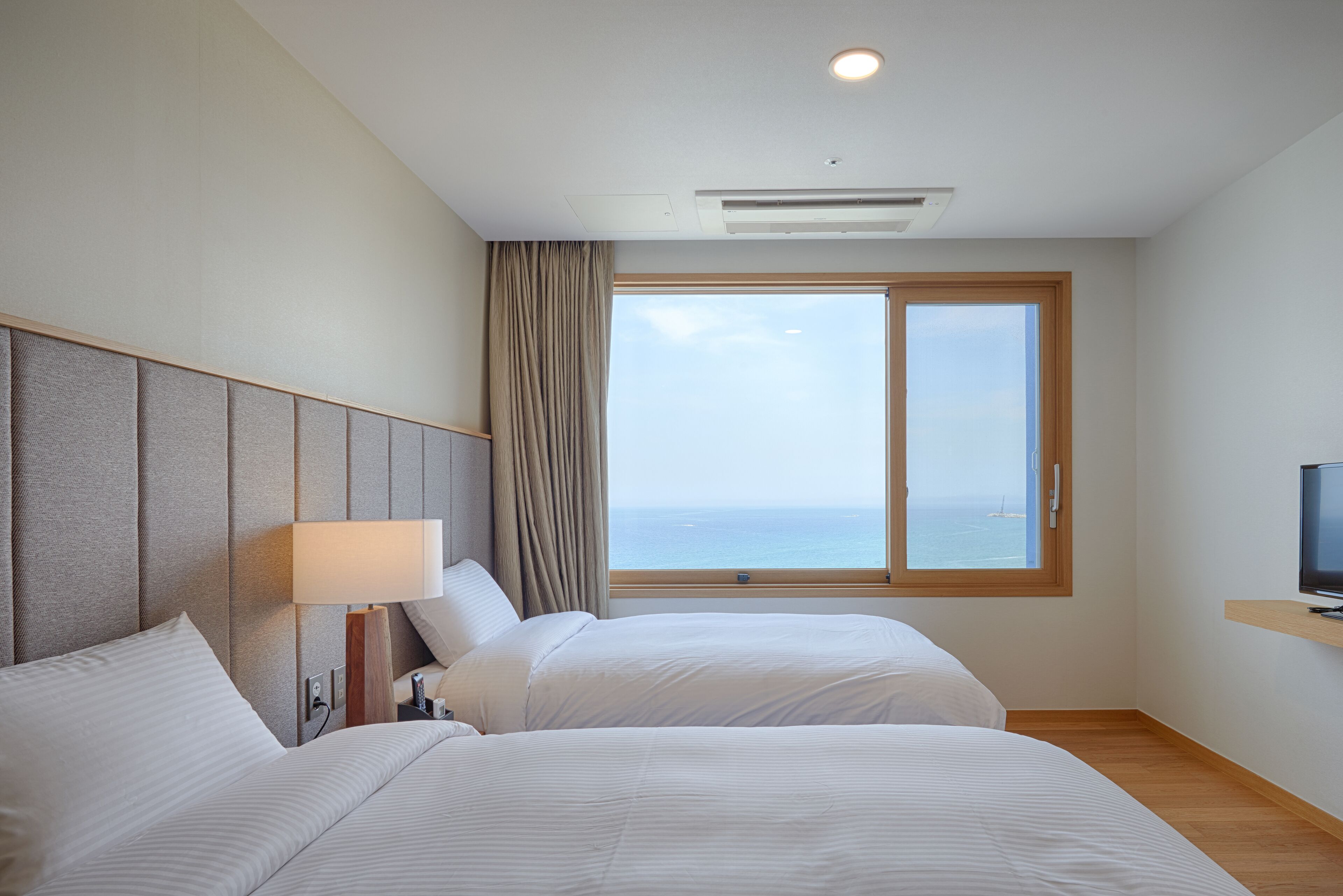 Habitación de hotel con dos camas, un ventanal con vista al mar, un televisor de pantalla plana sobre un estante de madera y un respiradero en el techo.