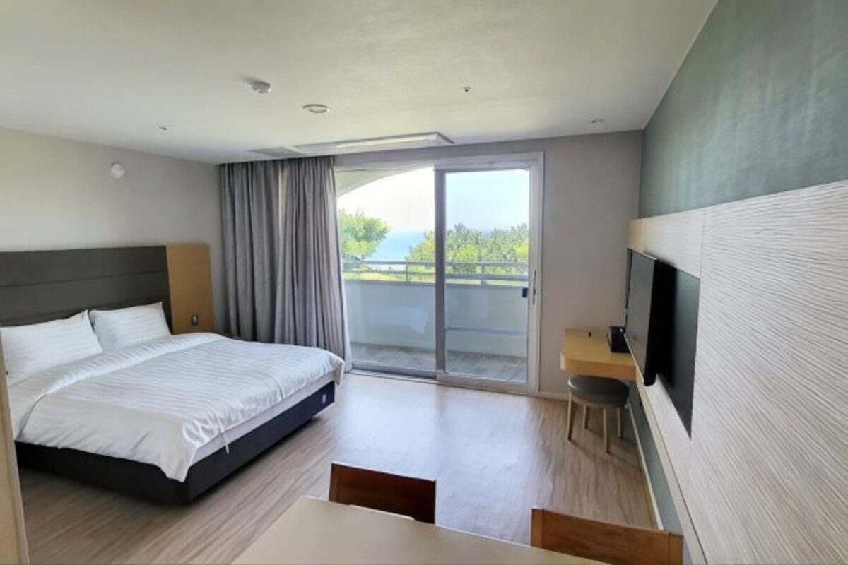 Habitación de hotel con cama, escritorio, silla, televisor y ventana corrediza al exterior.