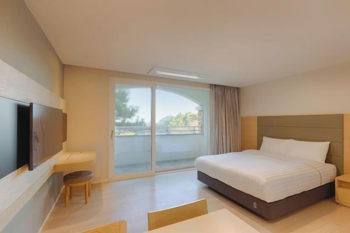 A modern hotel room with a large bed, a flat-screen TV, a small table, and a sliding glass door to the outside.