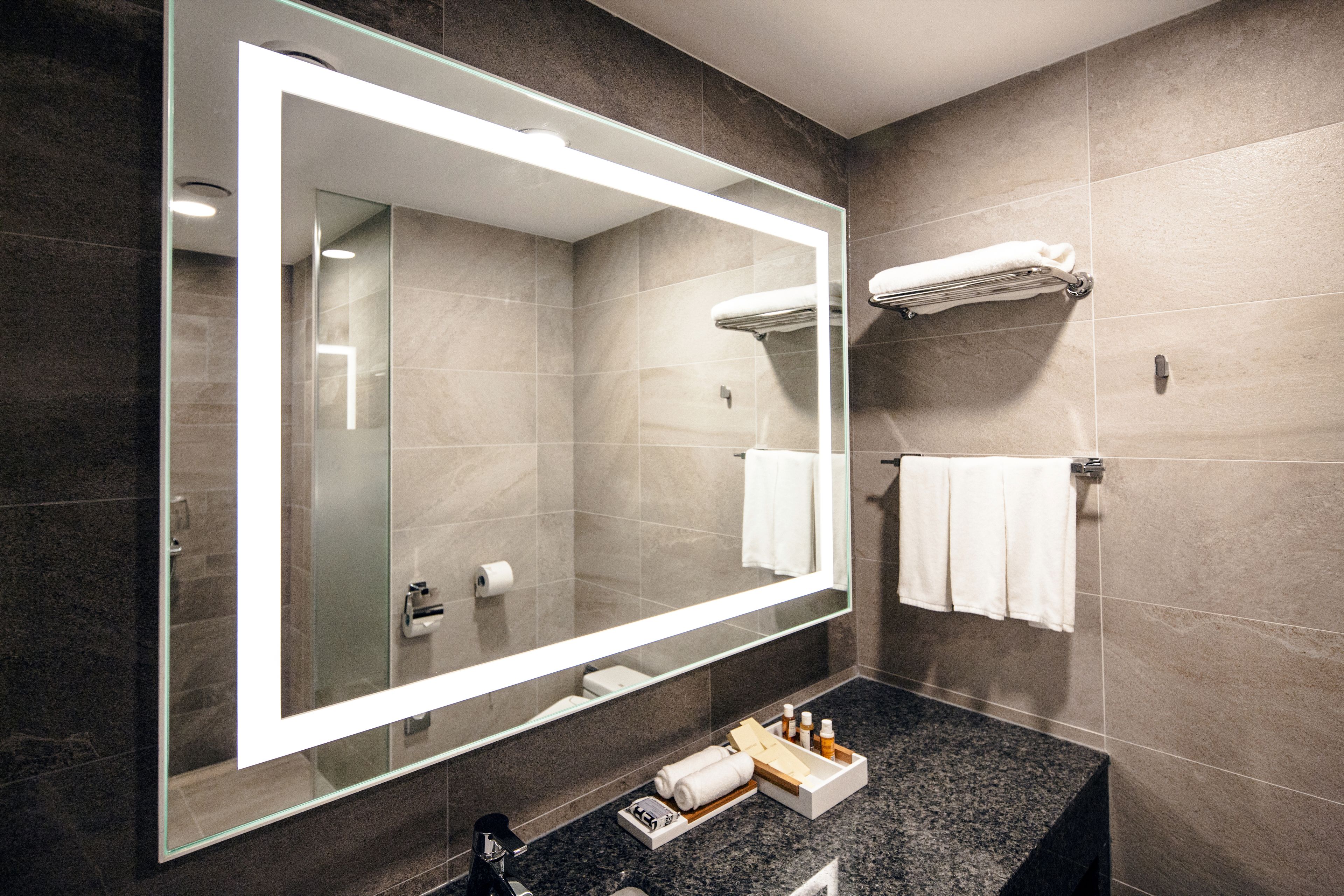 A modern bathroom with a large mirror, dark tiles, a towel rack, and a vanity with toiletries.