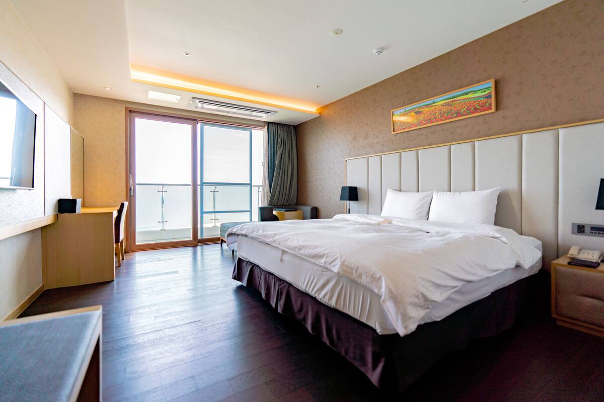 A hotel room with a large bed, a desk, a chair, a bedside table with a lamp, a painting on the wall, and a sliding door to the balcony.