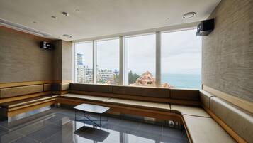 A modern hotel room with a large window, a seating area, and a flat-screen TV mounted on the wall.