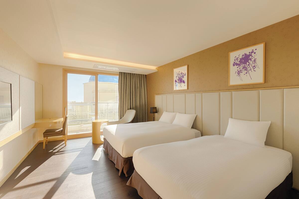 A hotel room with two beds, a desk, a chair, a window with a view, and two framed pictures on the wall.