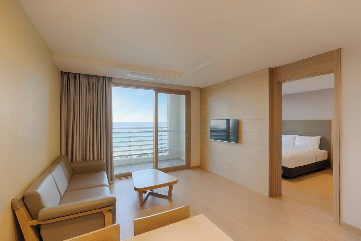 A modern hotel room with a sofa, a small table, a bed, and a large window offering a view of the sea.