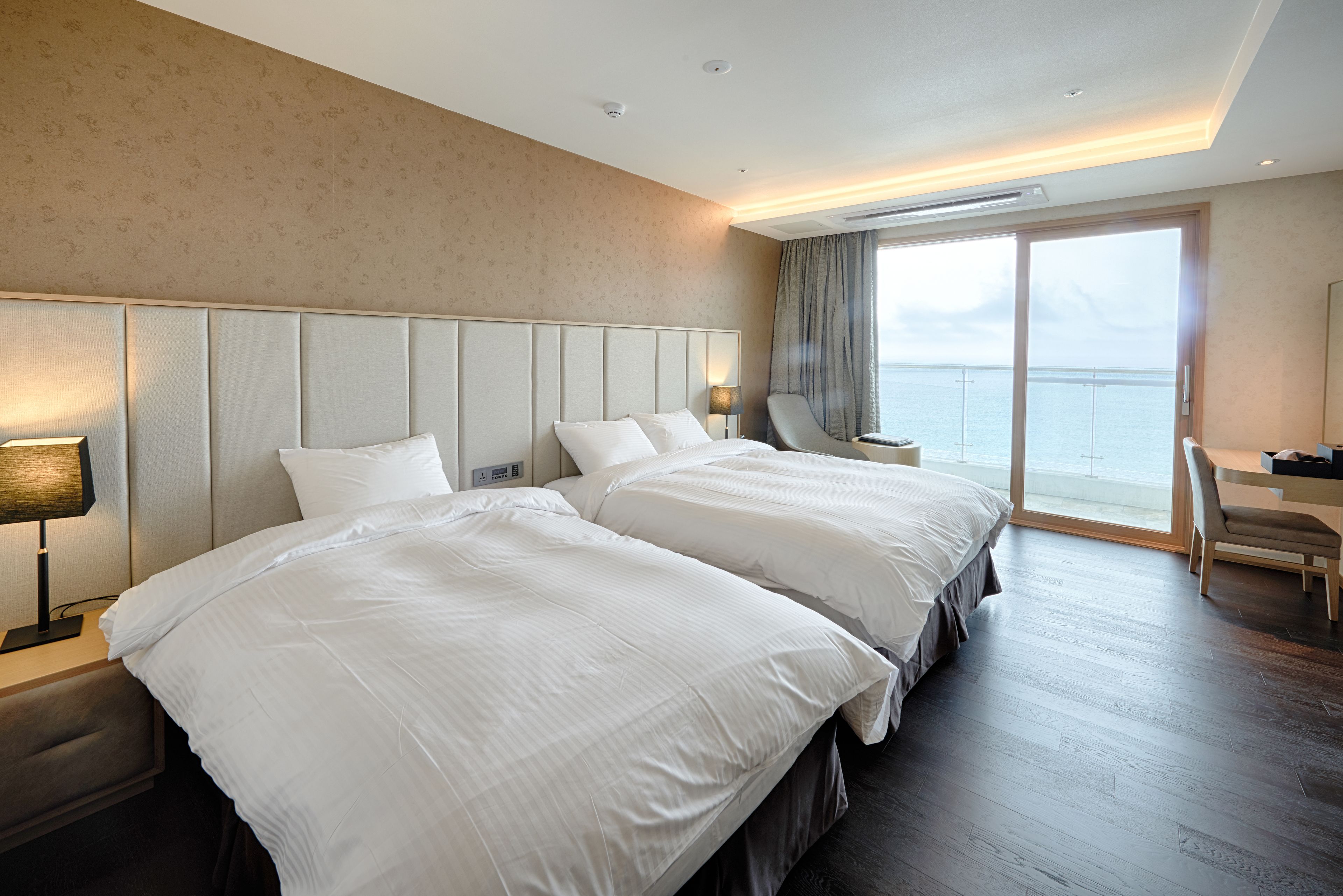 A hotel room with two beds, a desk, a chair, and a view of the sea.