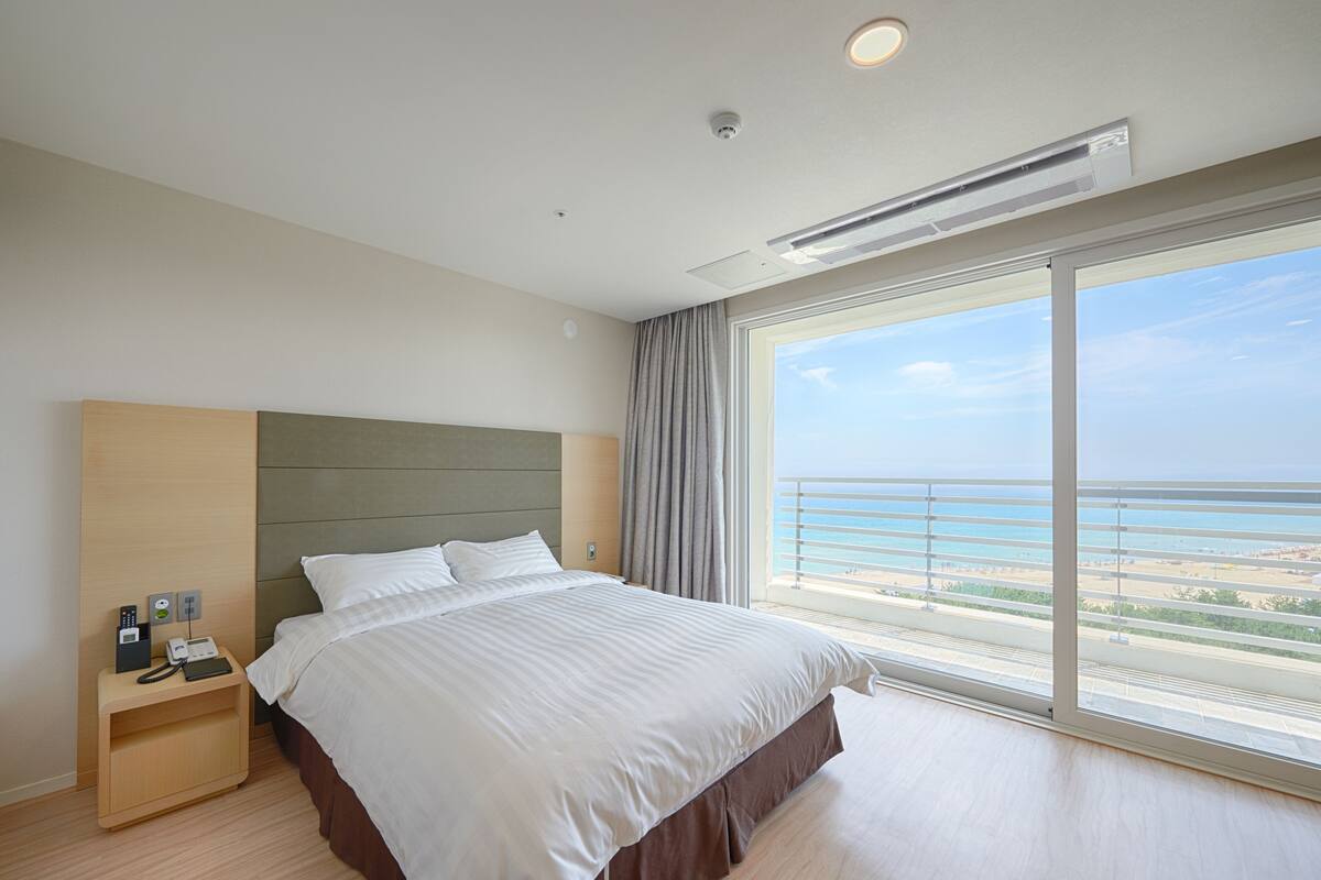 A hotel room with a large bed, a nightstand with a phone and a lamp, and a view of the ocean through a large window.
