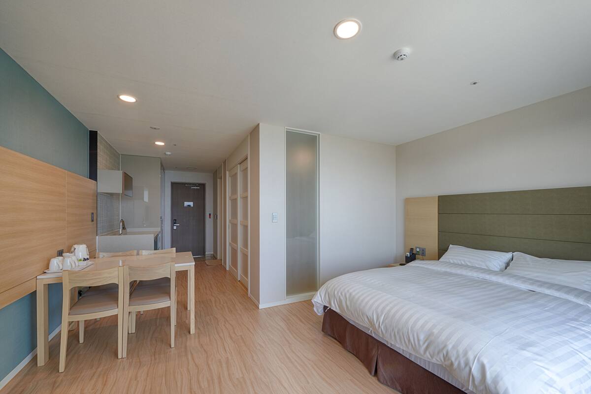 A modern hotel room with a wooden dining table, a bed with white linens, and a walk-in closet.