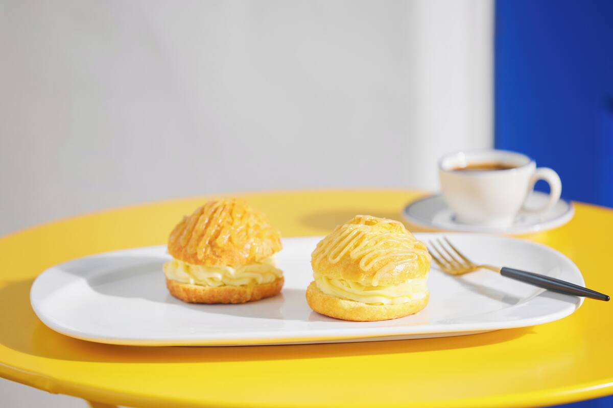 Two cream puffs on a white plate with a fork, placed on a yellow table with a white cup of coffee in the background.