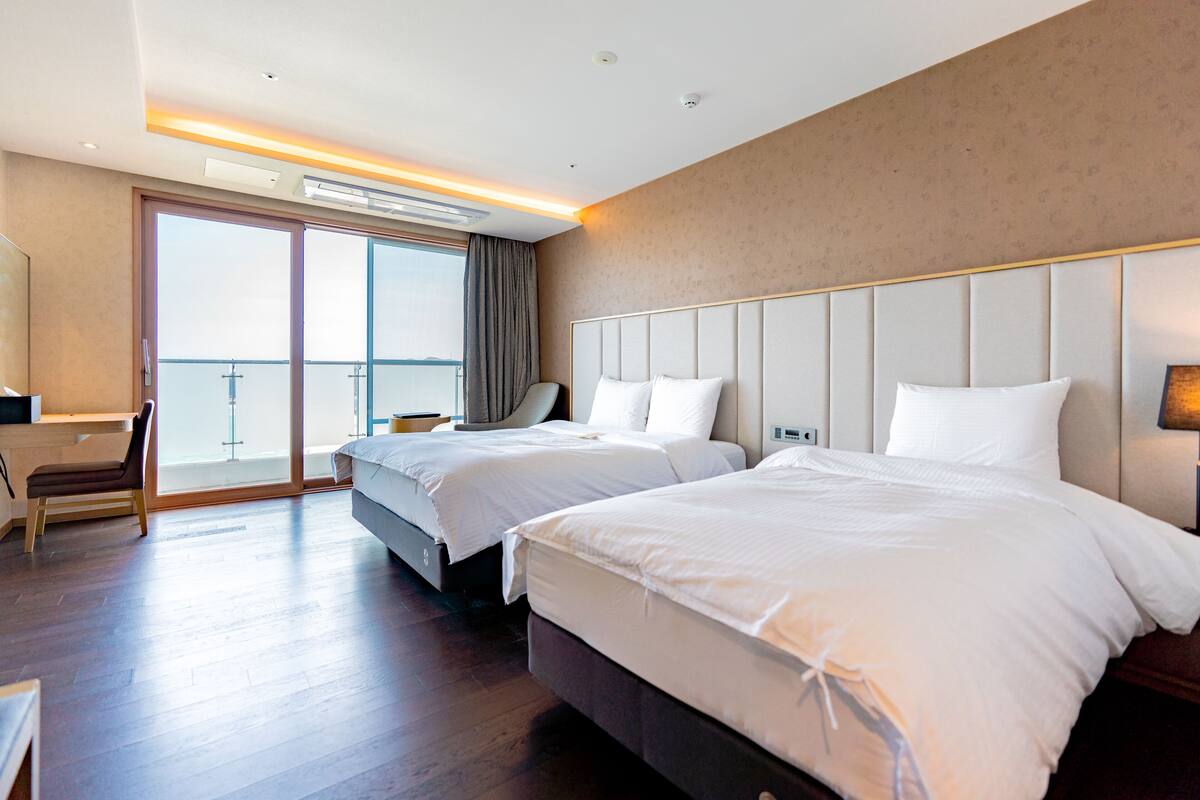A hotel room with two beds, a desk, and a chair. There is a large window with a view of the outside.