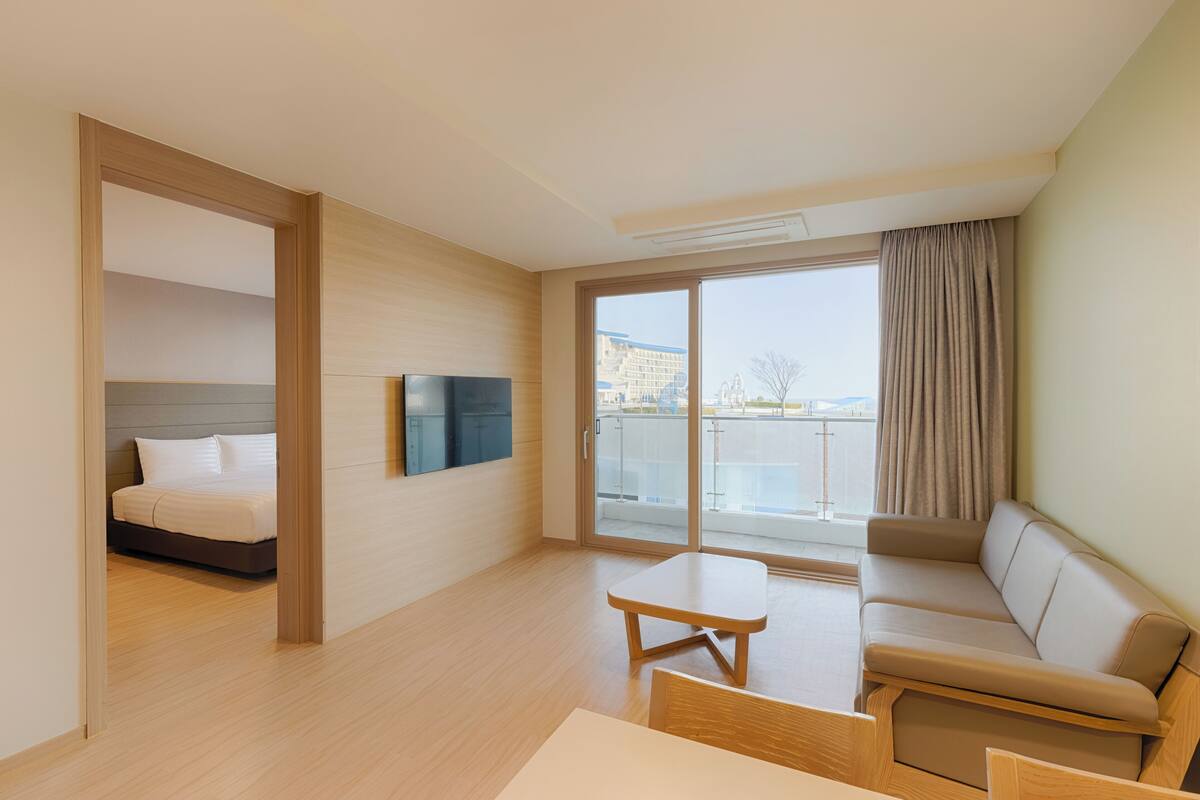A modern hotel room with a bed, a sofa, a TV, and a balcony with a view of the city.