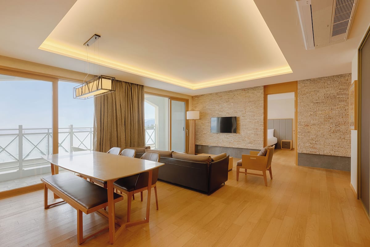 A modern hotel room with a wooden floor, a sofa, a dining table with benches, a TV mounted on the wall, and a large window with curtains.