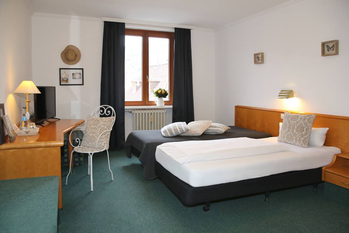 A hotel room with a bed, a desk with a lamp, a chair, and a window with curtains.