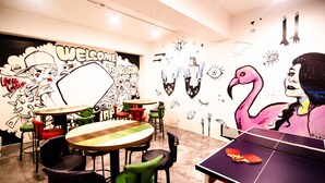 A room with a ping pong table, green and red chairs, and a wall mural with various characters and designs. - Kimchee Busan Downtown Guesthouse - Hostel (Busan)