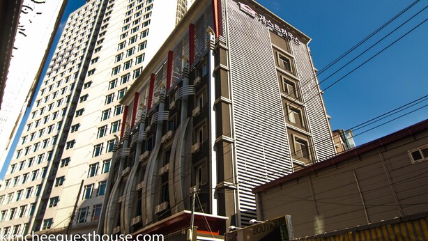 A multi-story building with a modern architectural design, featuring a mix of vertical and horizontal lines, and a prominent entrance with a red awning. - Kimchee Busan Downtown Guesthouse - Hostel (Busan)