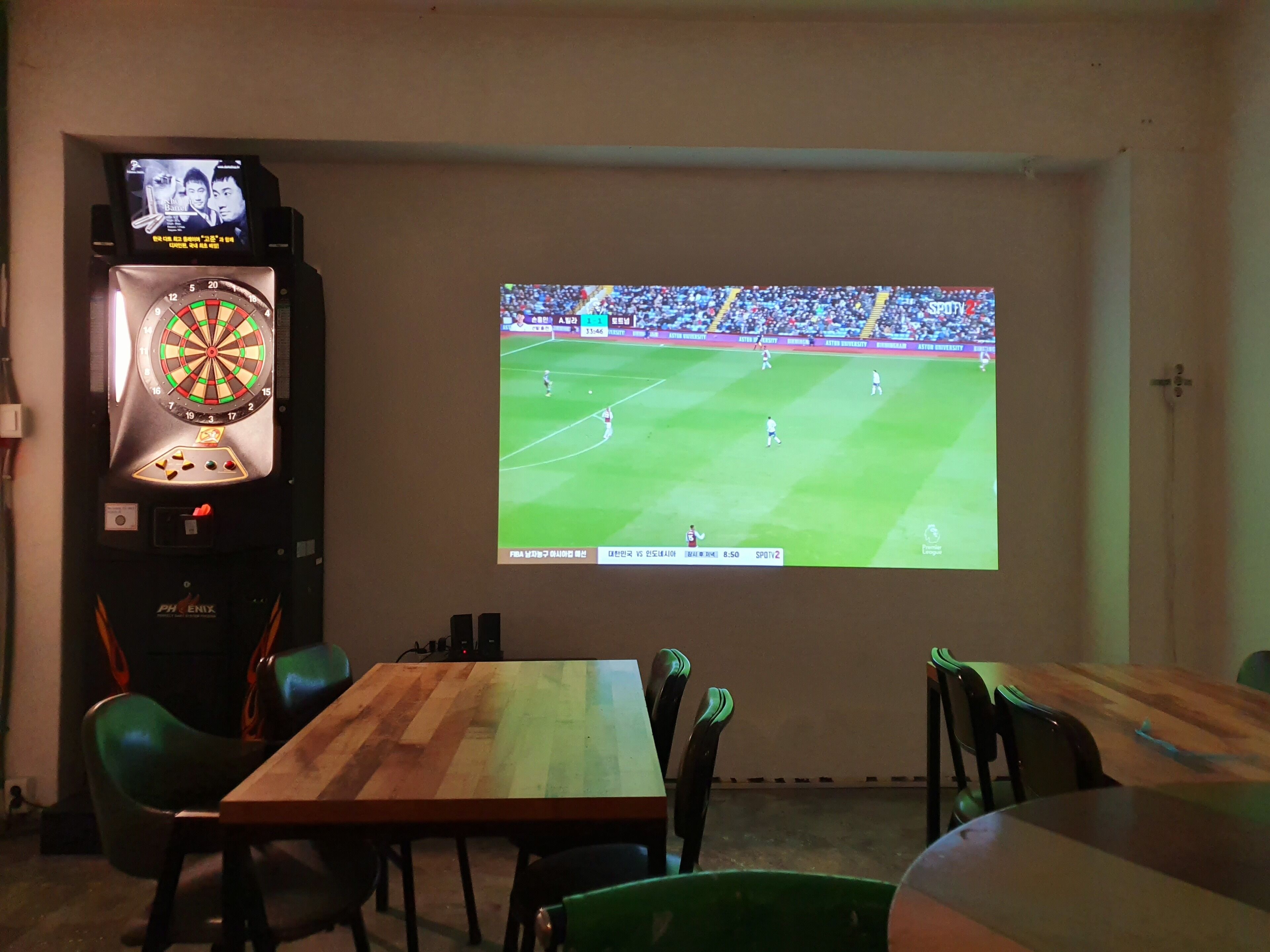 Een kamer met een dartbord, een televisie waarop een voetbalwedstrijd wordt vertoond, en een houten tafel met stoelen.