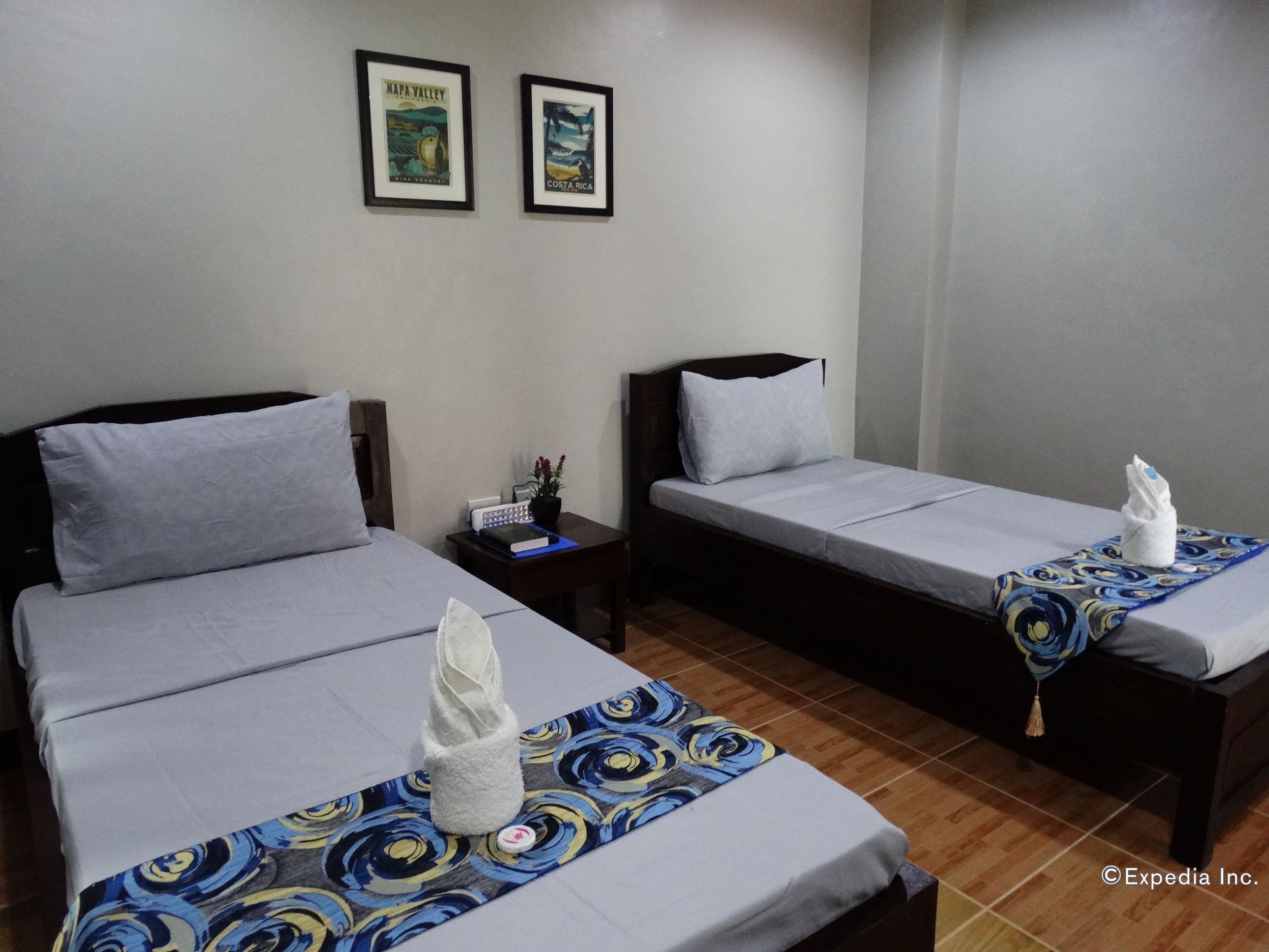 A hotel room with two single beds, each with a pillow and a towel. There is a small table with a book and a plant. Two framed pictures are on the wall.