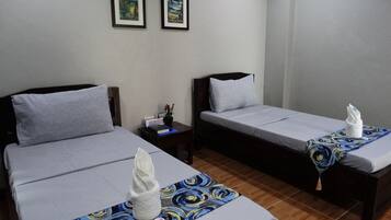 A hotel room with two single beds, each with a pillow and a towel. There is a small table with a book and a plant. Two framed pictures are on the wall.