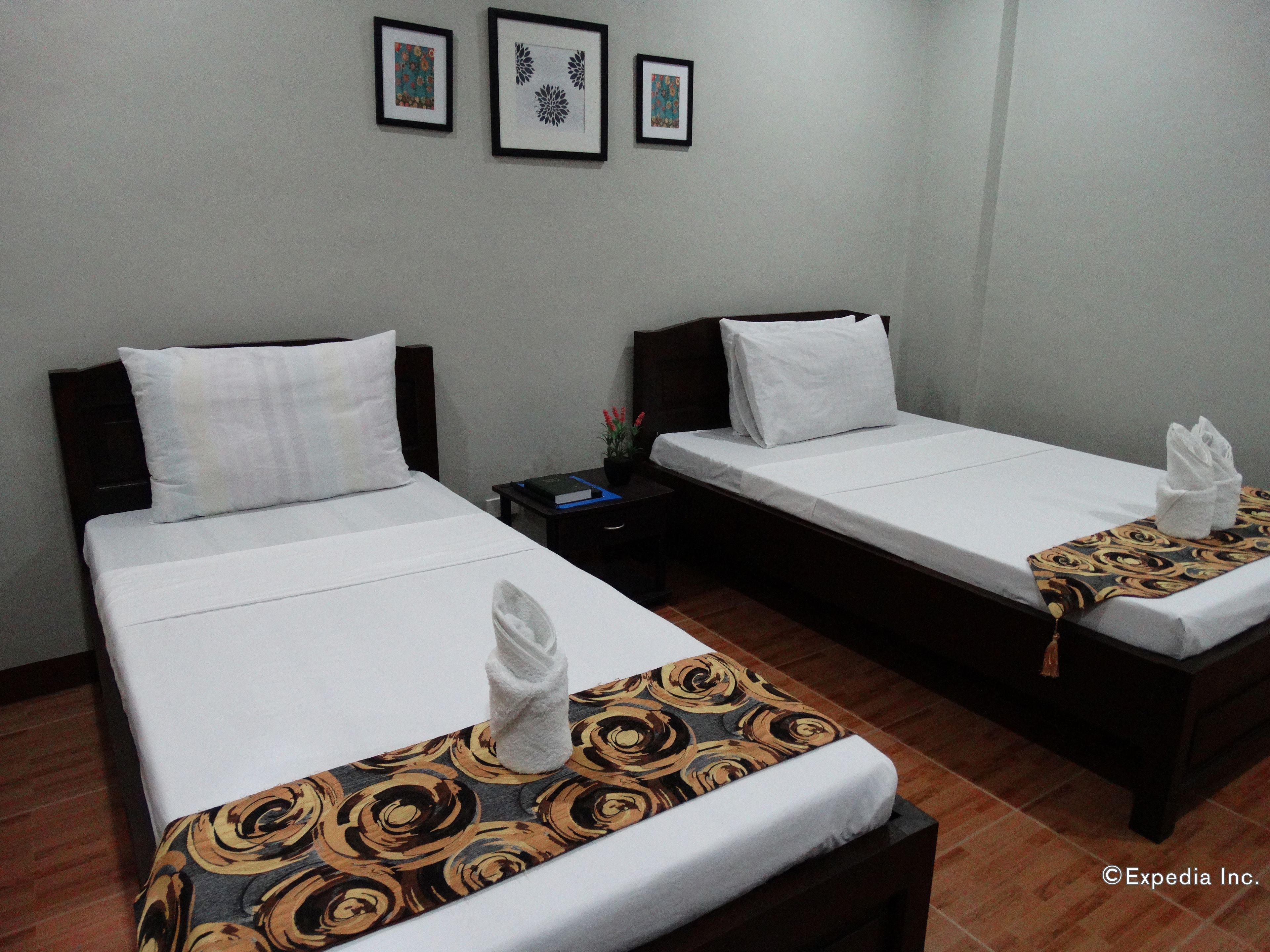 A hotel room with two single beds, each with white linens and a patterned bed runner. There are framed pictures on the wall above the beds.