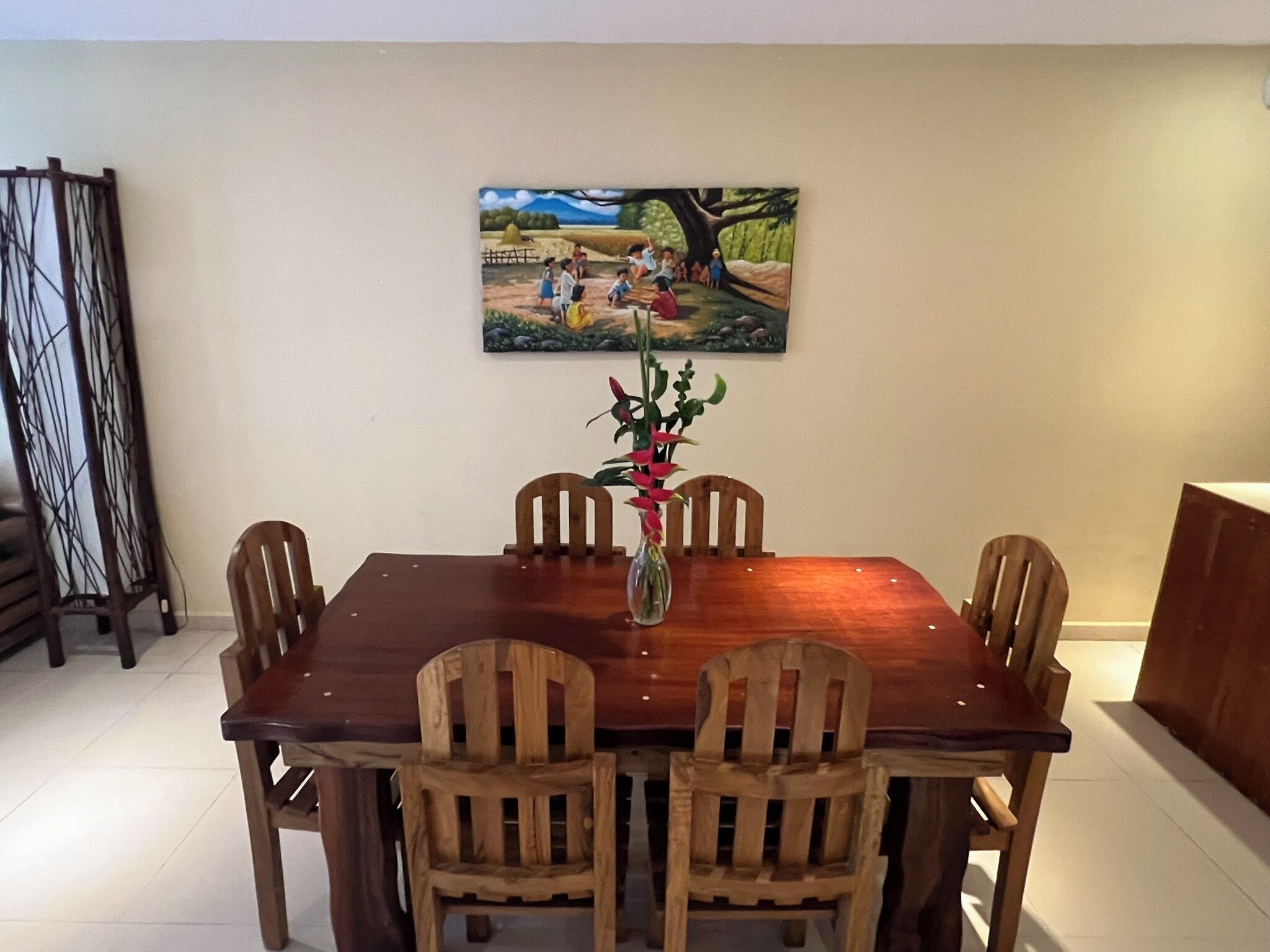 A dining area with a wooden table and chairs, a painting on the wall, and a vase with flowers.
