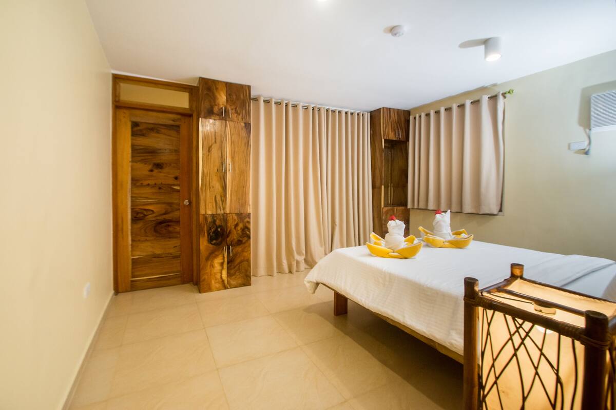 Une chambre d’hôtel avec un lit, une porte en bois, des rideaux et une petite table sur laquelle se trouvent des fruits.