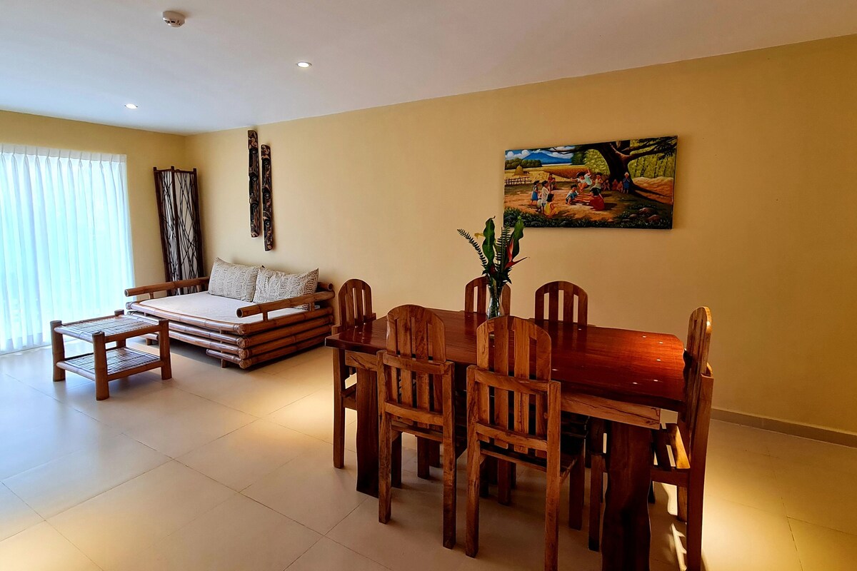 Un salón con sofá, mesa de comedor de madera y sillas. Hay un cuadro en la pared y una ventana con cortinas.