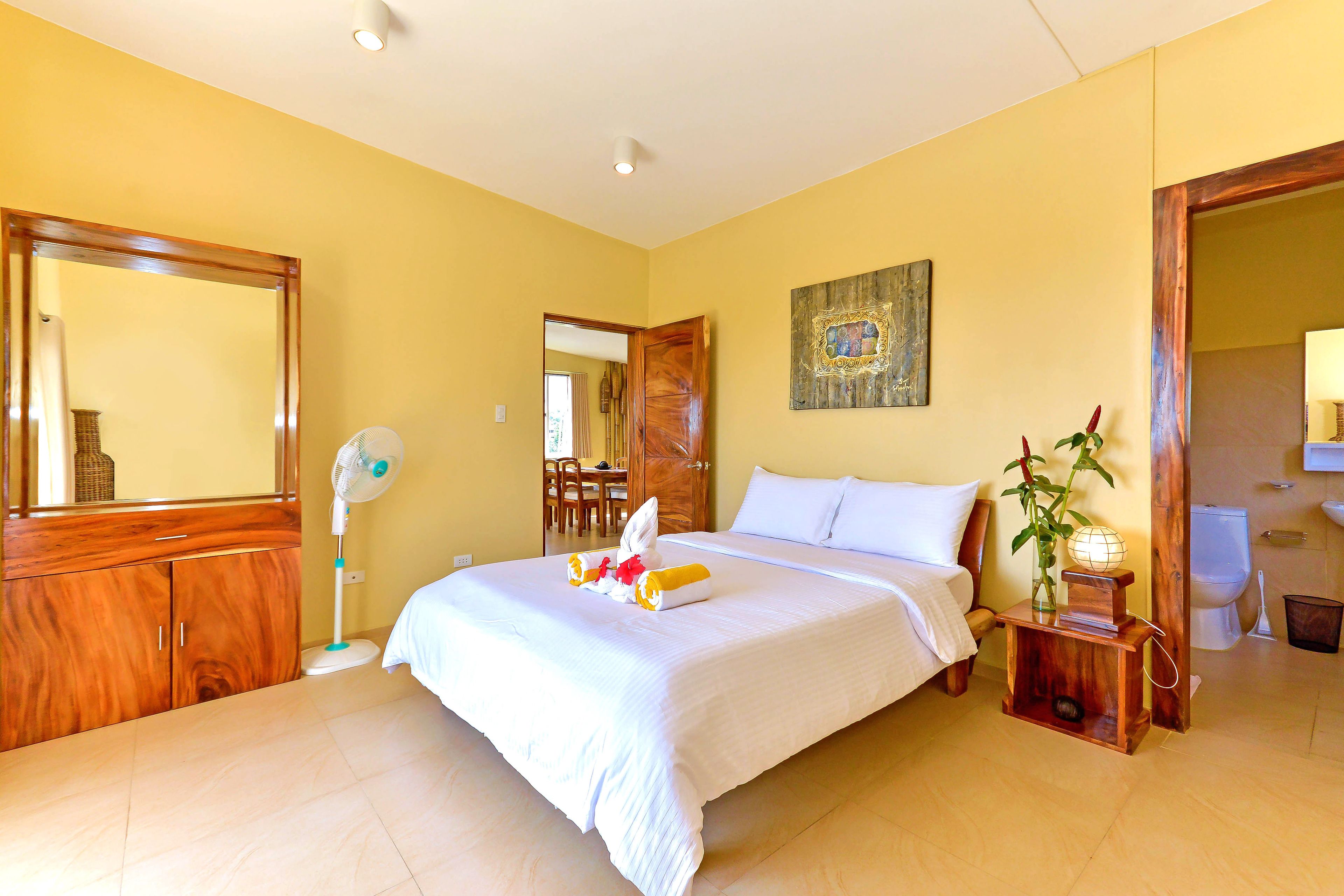 Une chambre d’hôtel avec un grand lit, une armoire en bois, une table de chevet avec une lampe, et une salle de bain visible à travers une porte ouverte.