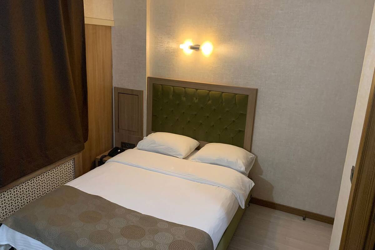 A hotel room with a bed, a headboard, a wall-mounted light fixture, and a curtain.