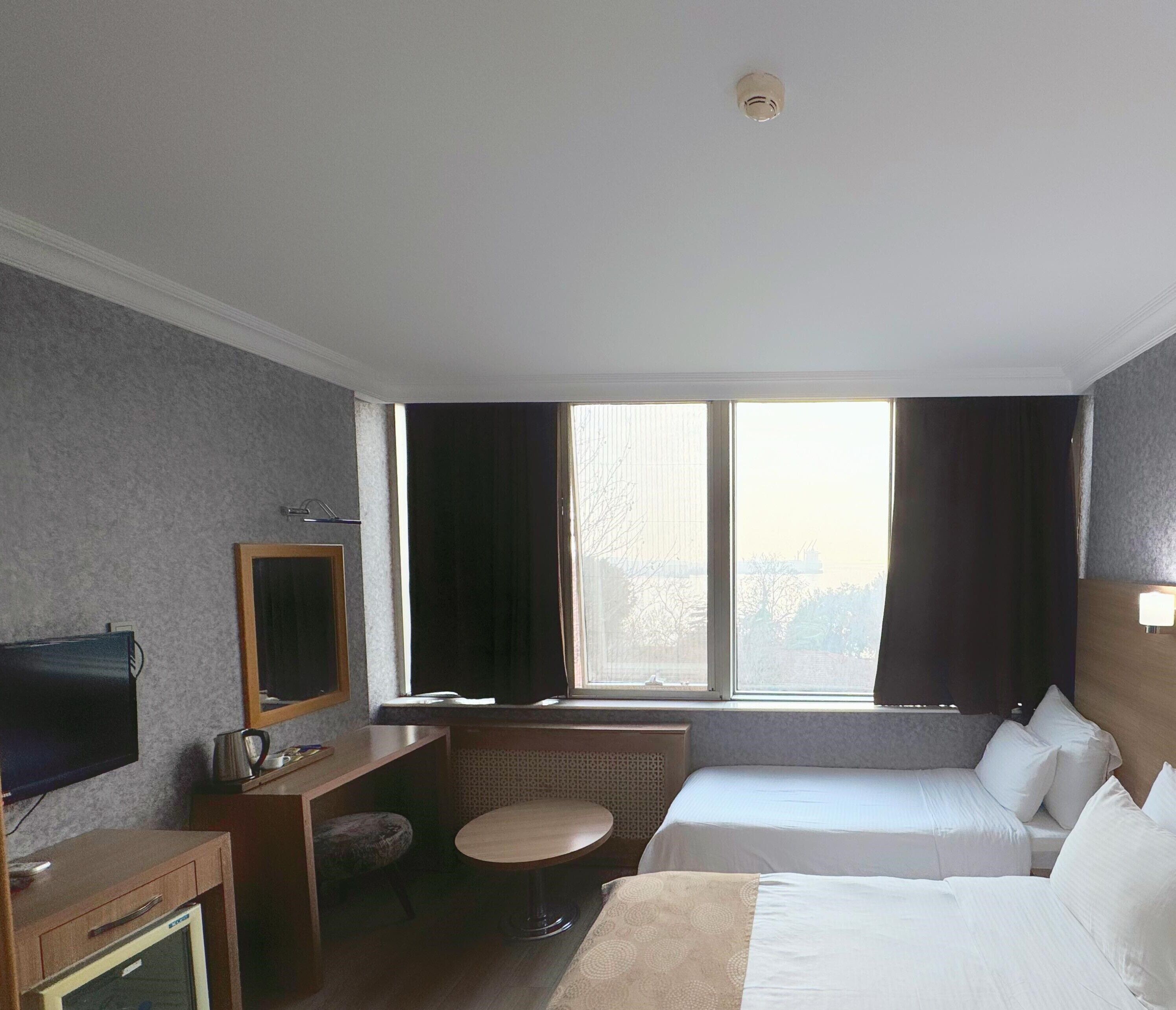 Habitación de hotel con cama, escritorio con silla, televisor, ventana con cortinas y vista a la ciudad.