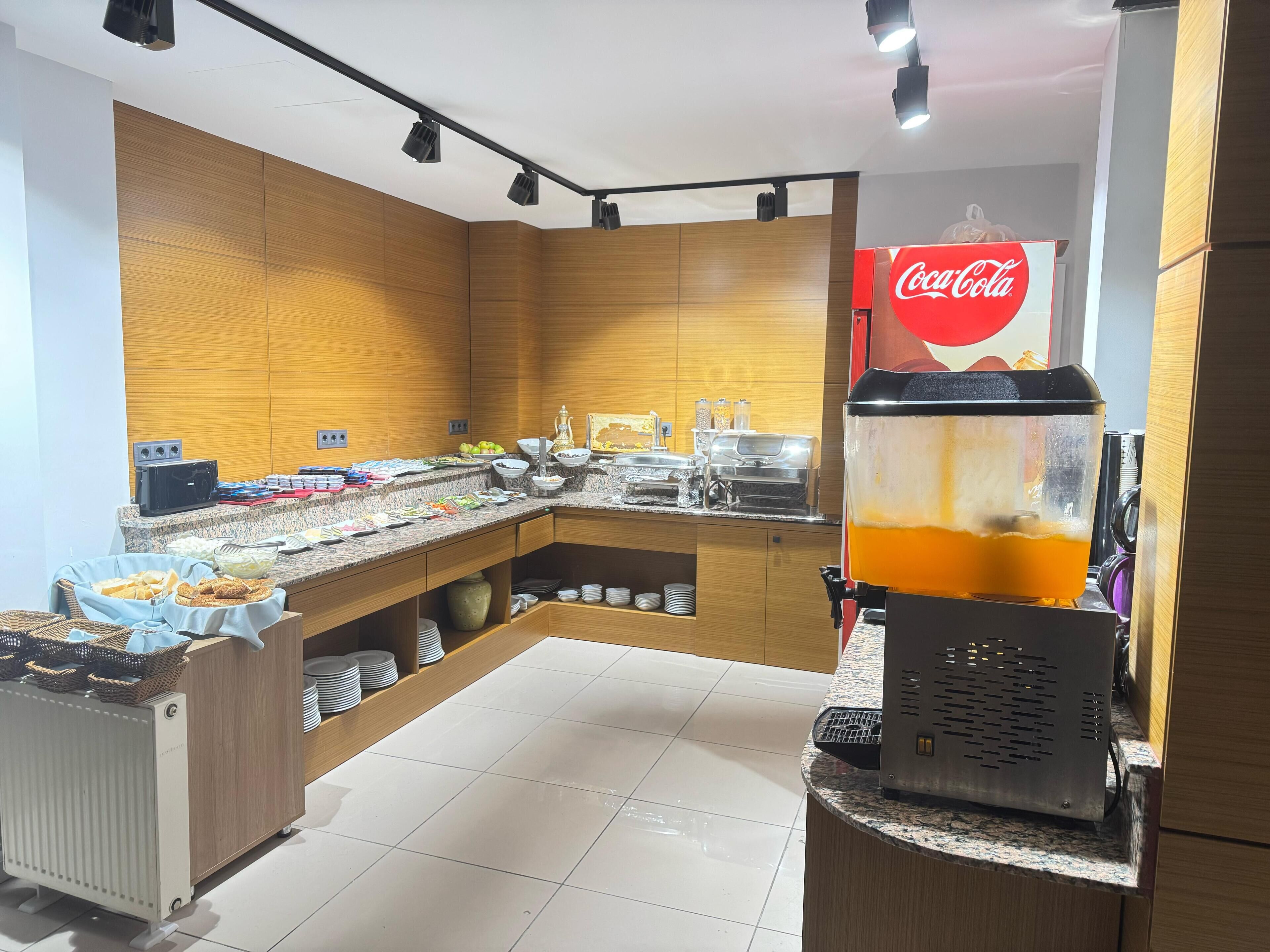 A modern kitchen with a buffet setup, including a Coca-Cola refrigerator, a juice dispenser, and various food items.