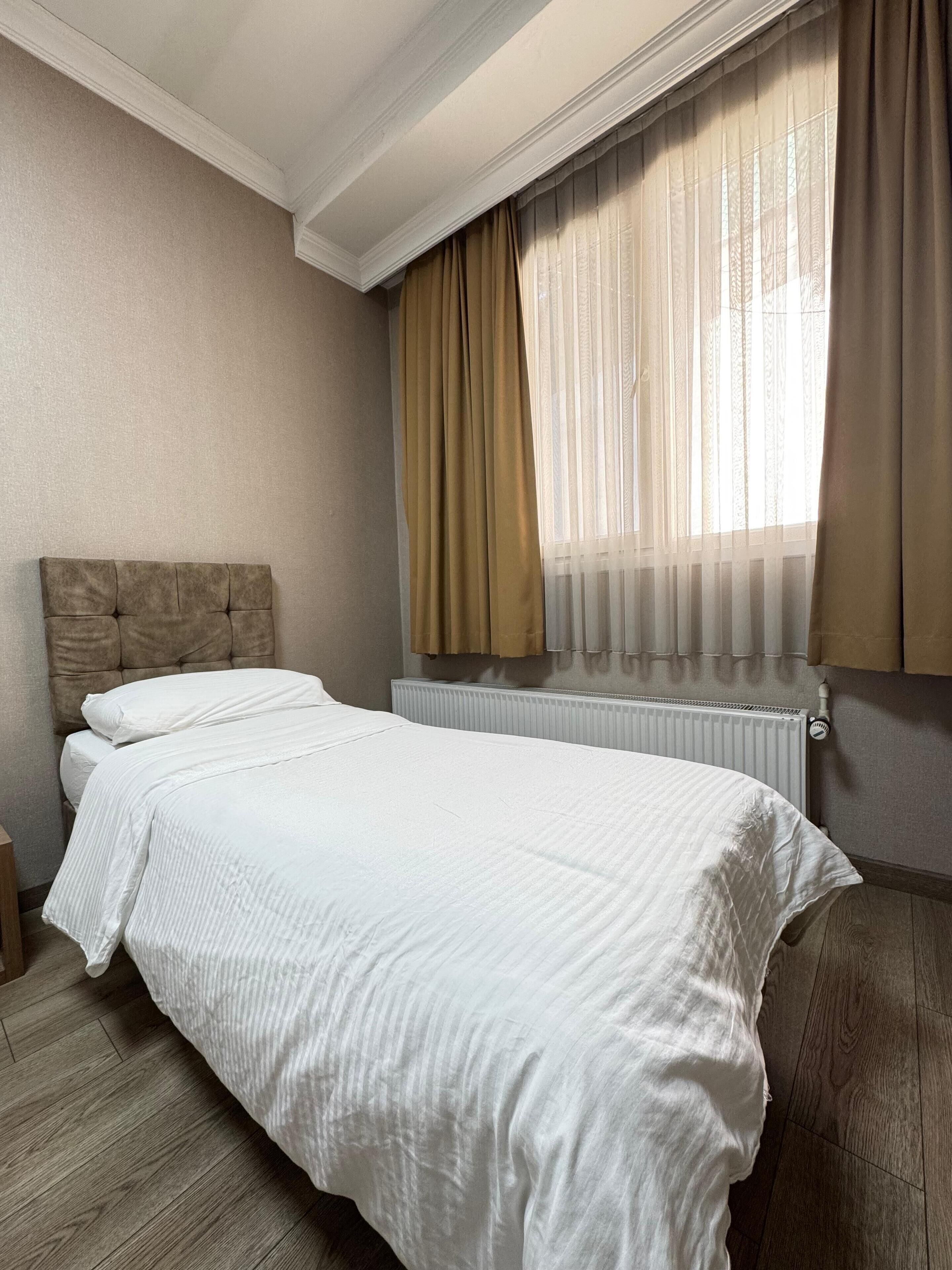 Una cama bien hecha con ropa blanca, cabecera y una ventana con cortinas traslúcidas.